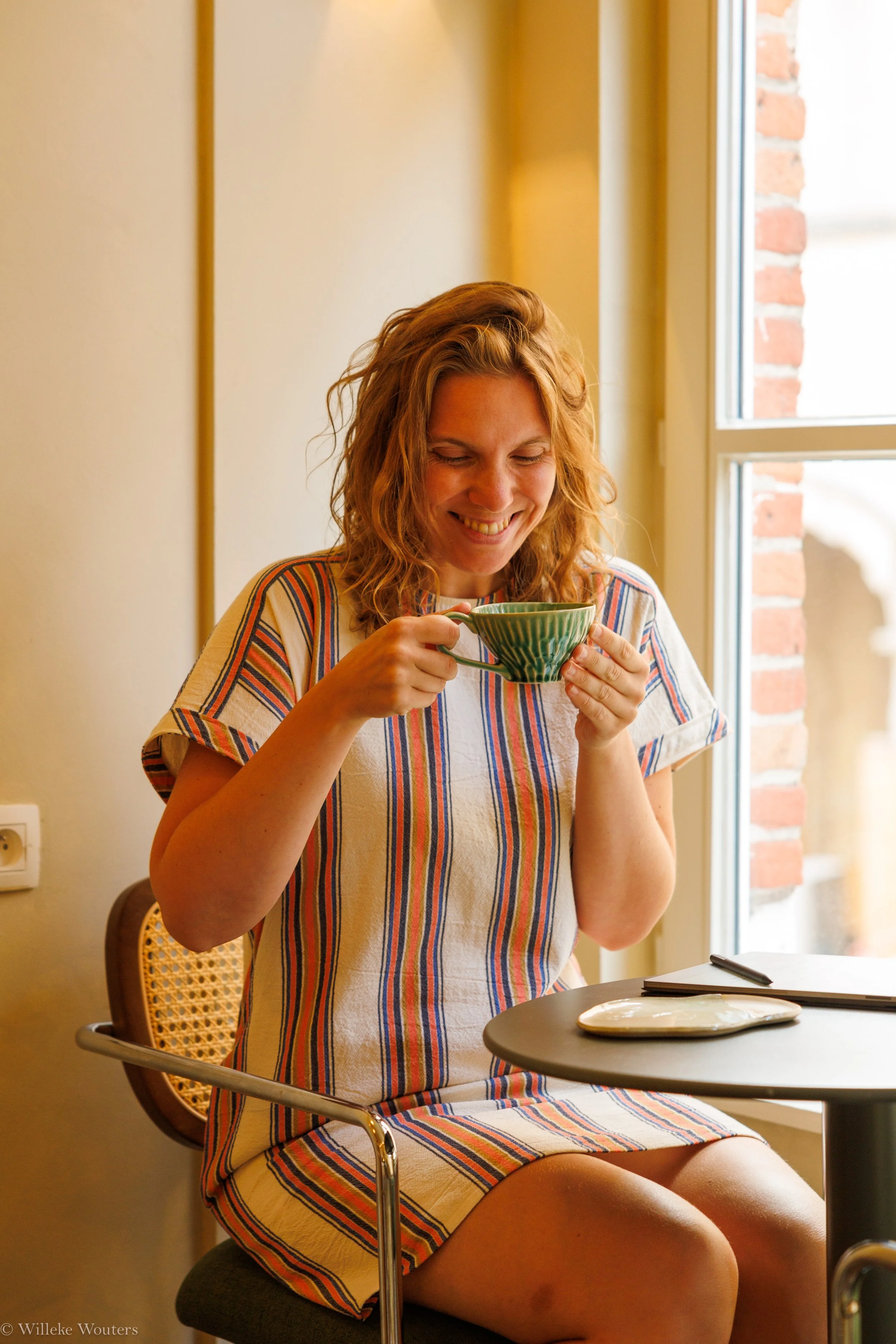 Vrouw in een kleurrijk gestreept jurk die lacht terwijl ze uit een groene kop drinkt, zittend aan een tafel in een kamer met een raam en bakstenen muur.
