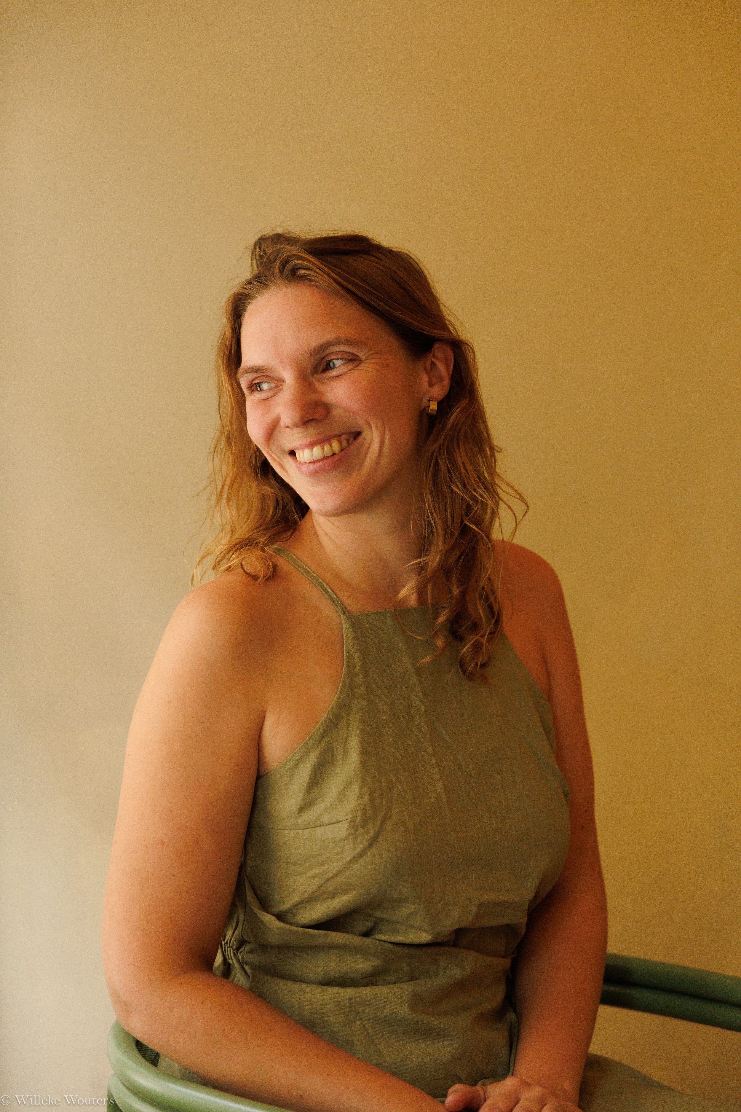 Een glimlachende vrouw met lang, golvend, kastanjebruin haar draagt een groen satijnen top en zit op een groene stoel, voor een lichte muur.