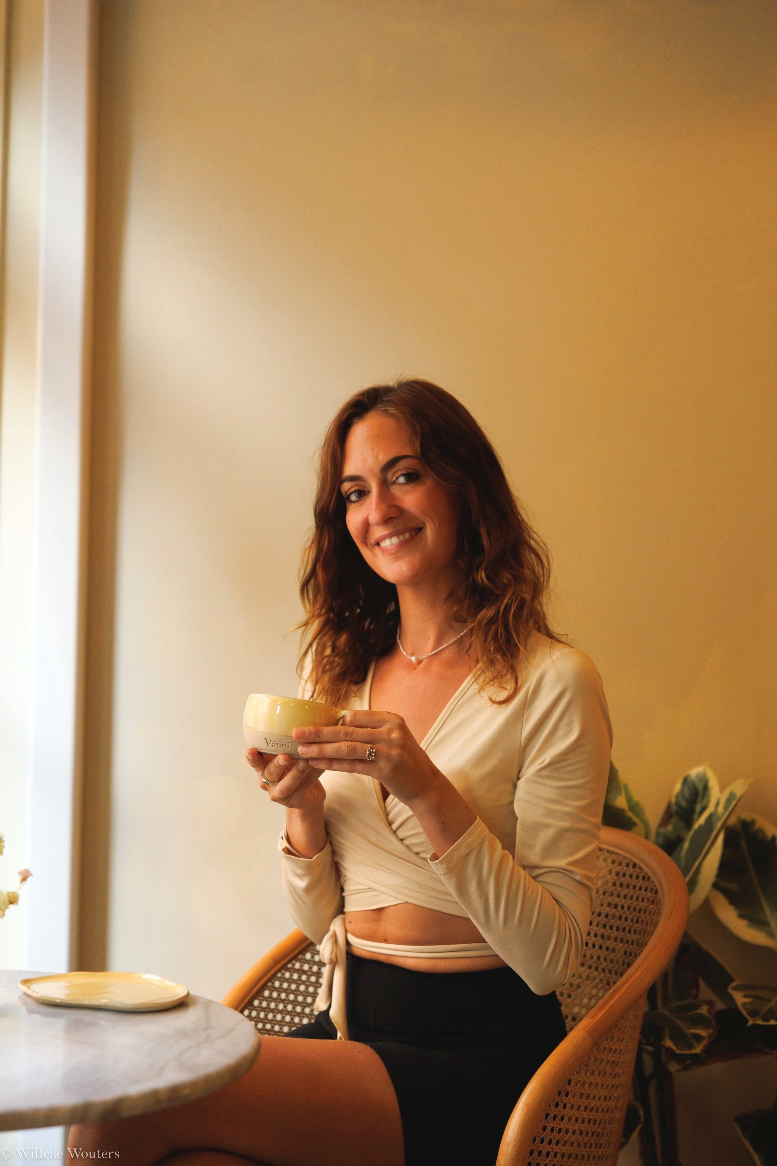 Een lachende vrouw met bruin haar die in een rustige binnenruimte staat en een kopje vasthoudt. Ze draagt een creme-gekleurde top en een zwarte rok en zit op een rieten stoel. Er zijn planten en een tafeltje met een bordje in de achtergrond.