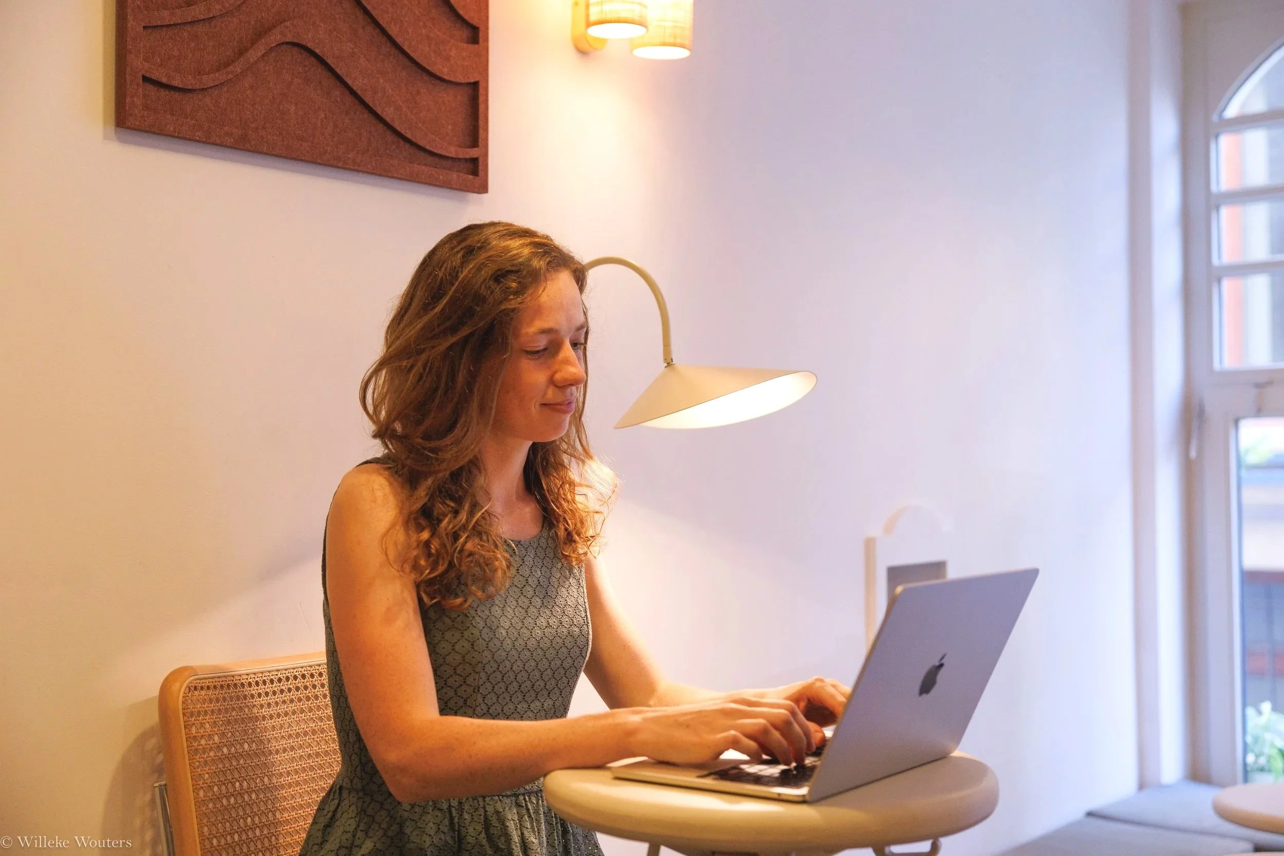 Vrouw met lang, ruw haar en een grijze jurk zit aan een ronde tafel ogenschijnlijk werkend op een laptop, binnen in een lichte kamer met grote ramen en wandverlichting.