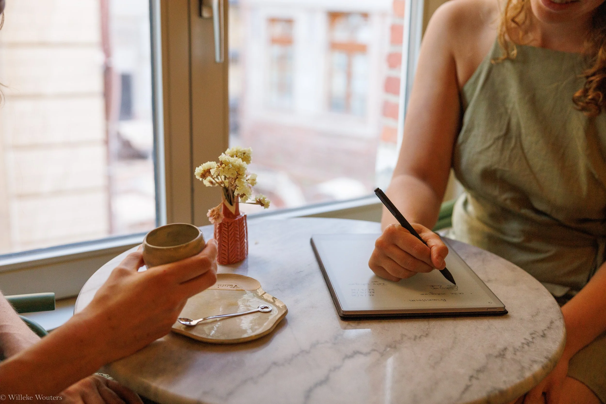 een persoon schrijft in een notitieboekje terwijl een andere hand een beker vasthoudt; er staat ook een vaas met bloemen op de tafel.