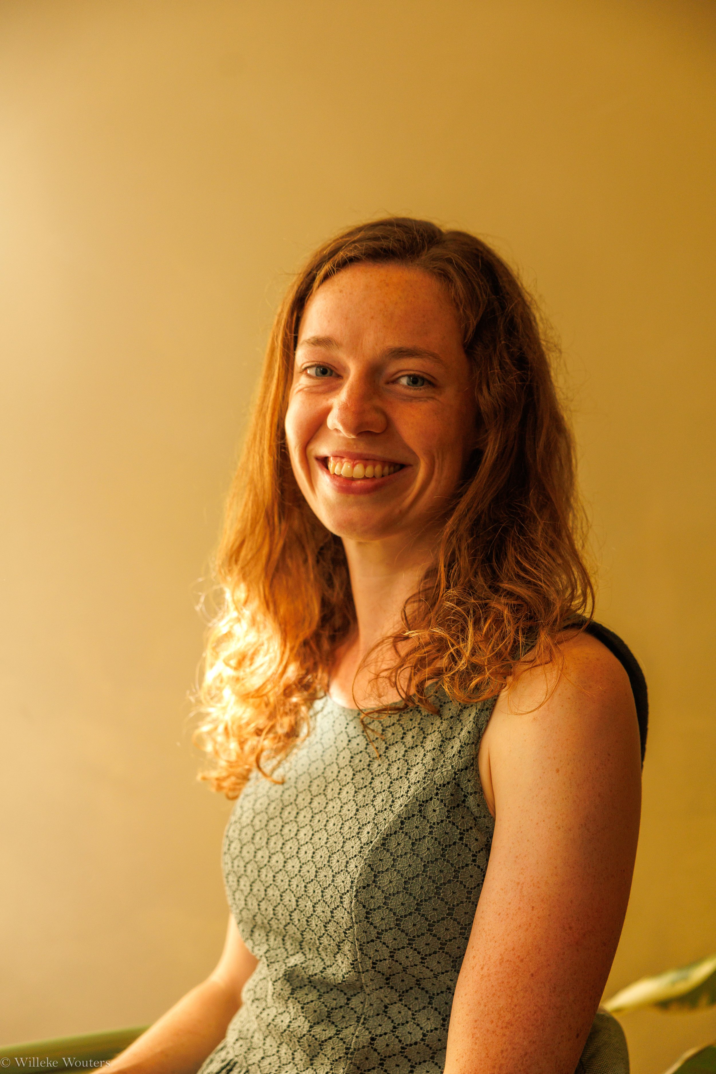 Portret van een jonge vrouw met rustiek krullend haar, glimlach, in een groene jurk met een patroon, zittend in een kamer met een gelijkkleurige muur.