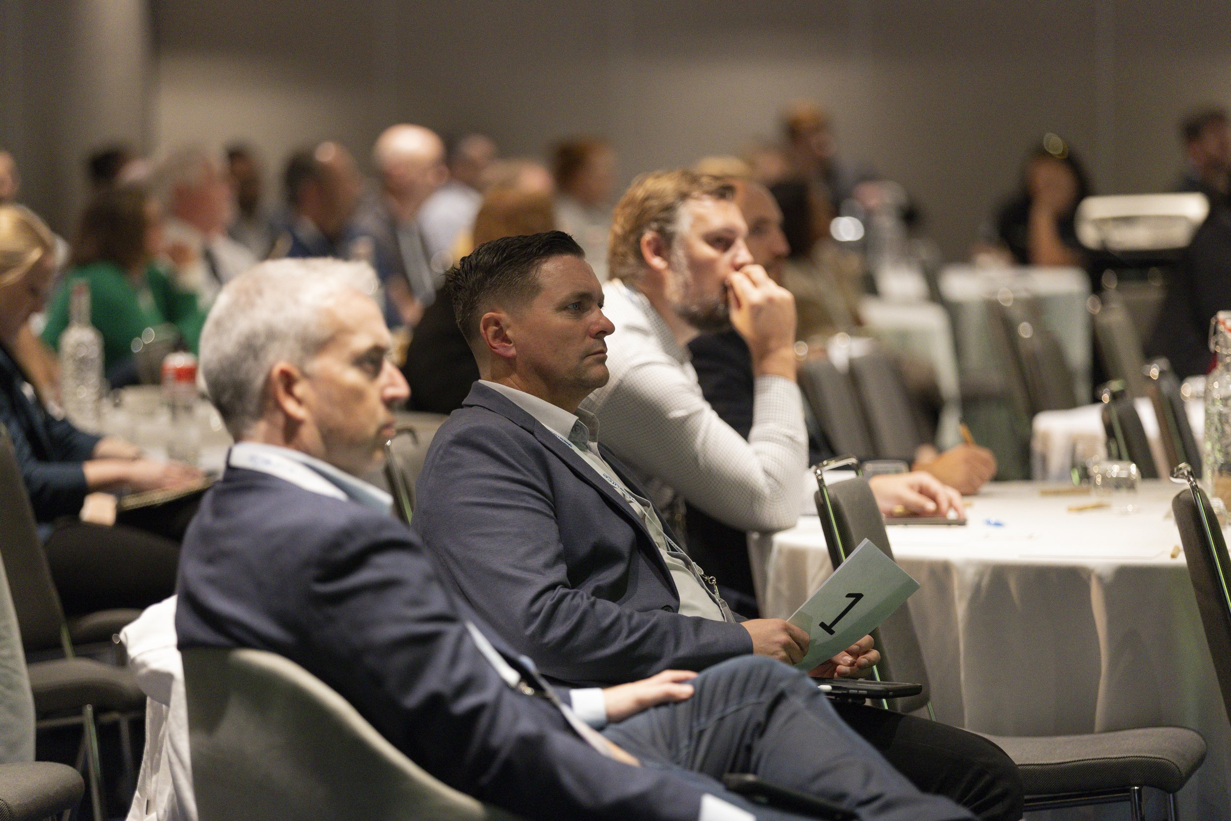 Conference attendees seated at tables listening to a presentation, with some holding papers or notepads.