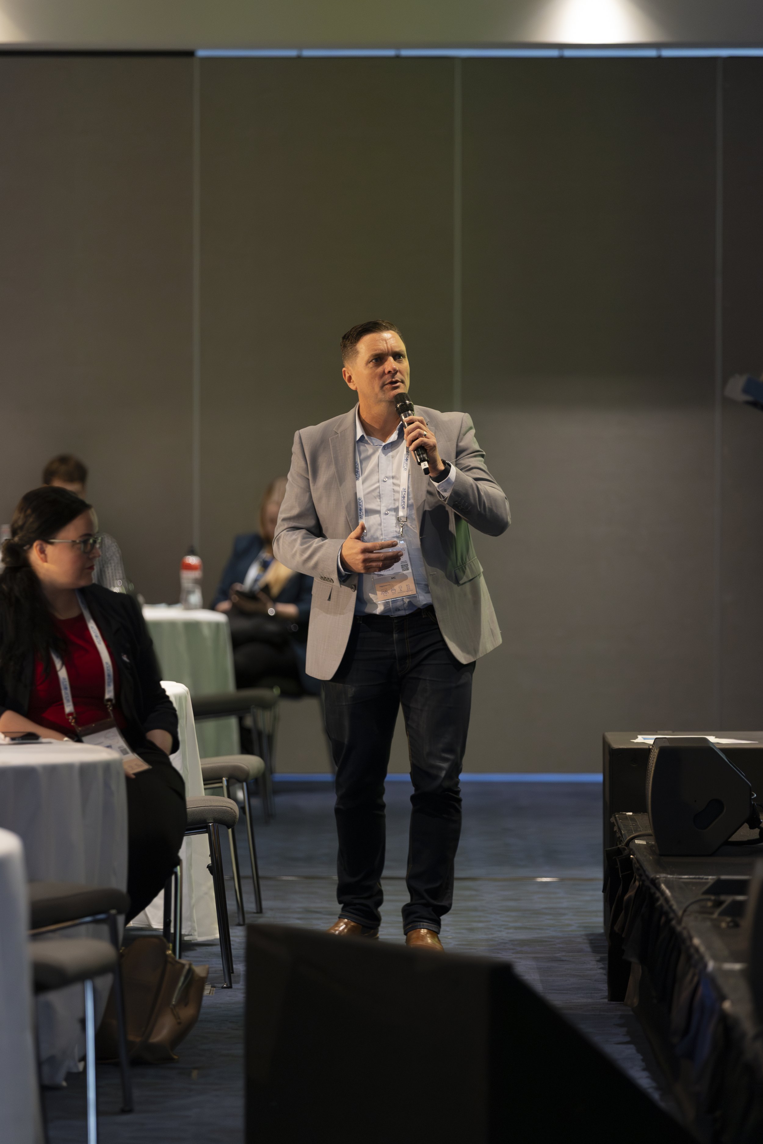 Man in gray suit speaking into a microphone at a conference or seminar, with audience members seated at tables around him.