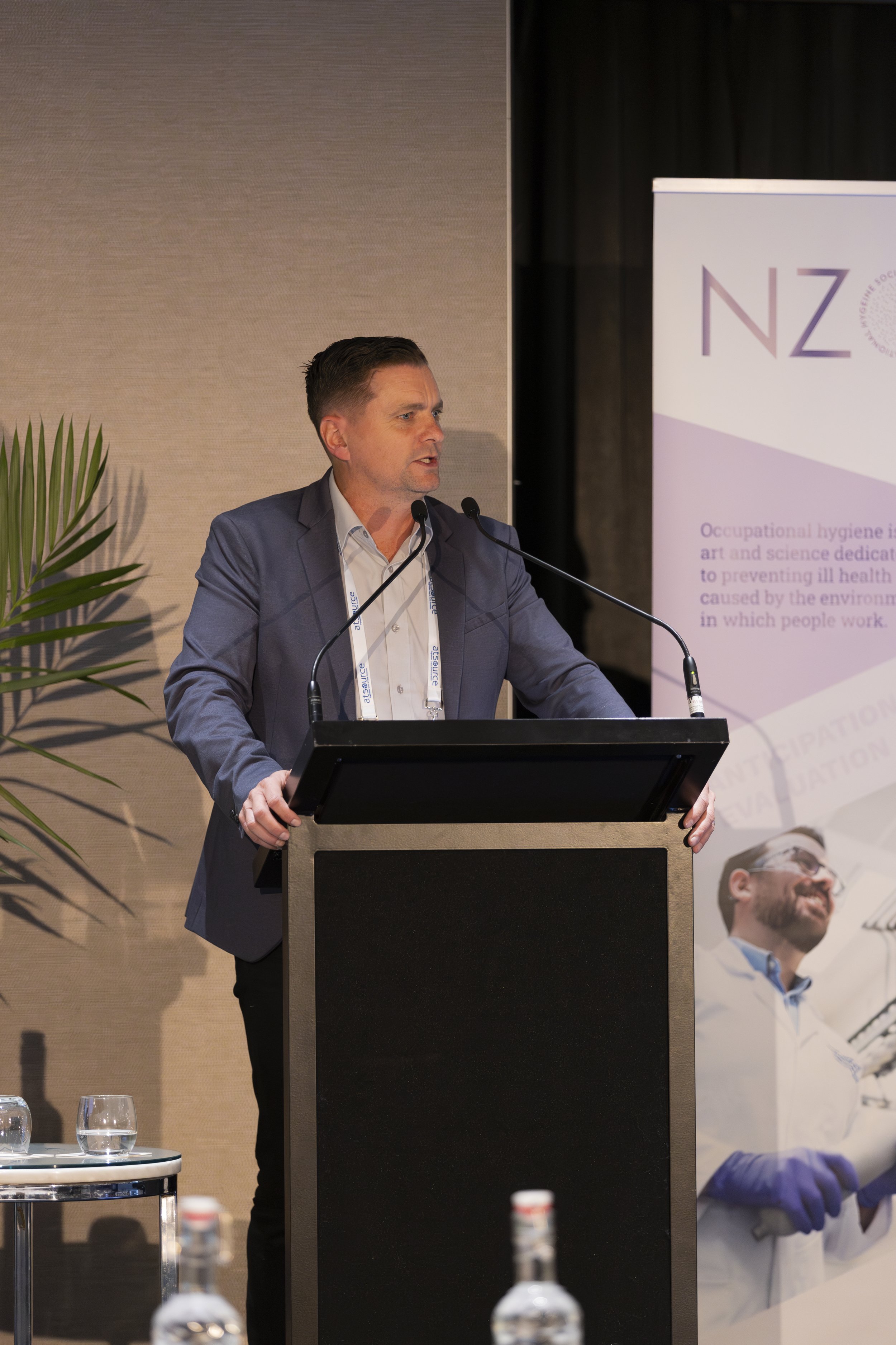 A man in a gray suit and white shirt is speaking at a podium during a conference or event. Behind him, there is a large banner with purple and pink text featuring the letter 'NZ' and additional text related to occupational hygiene and health topics. A woman in a white coat and purple gloves is also visible in the background, smiling and standing near the banner. There's a small round table with glasses of water in the foreground.