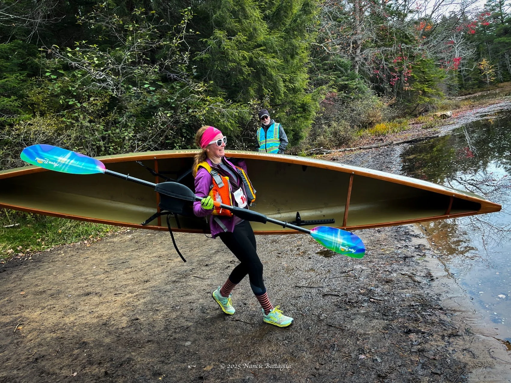A woman carrying a canoe and a paddle, standing near a body of water, with a man wearing a reflective vest in the background, surrounded by trees and autumn foliage.