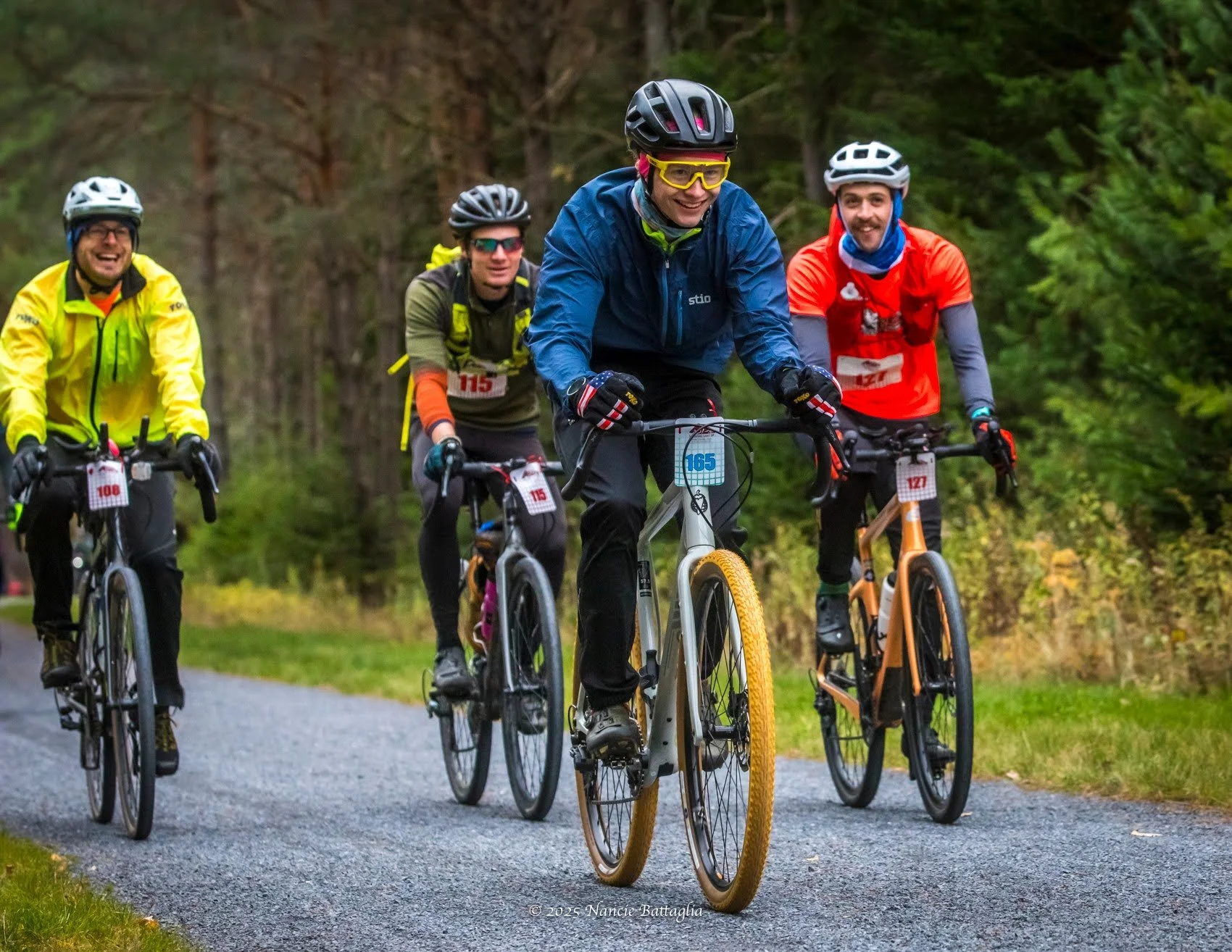 A group of five cyclists riding on a gravel trail in a wooded area, wearing helmets and colorful outdoor gear, with one cyclist leading and smiling.