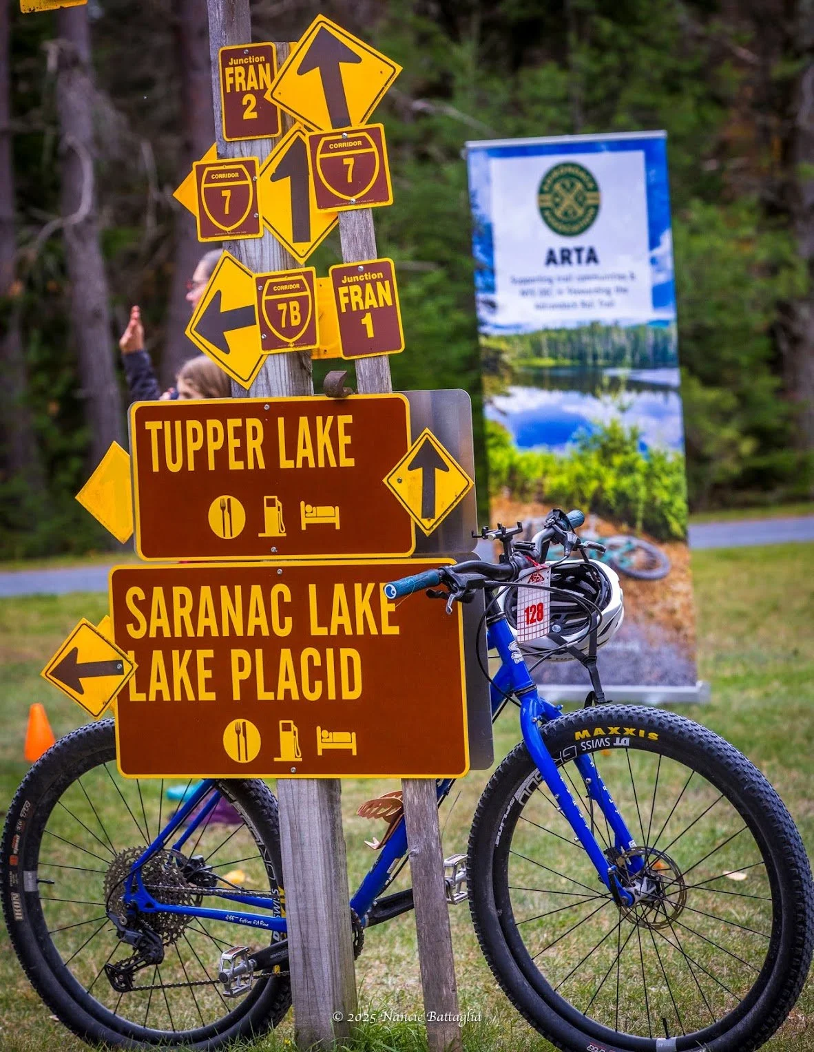 Signposts pointing to Tupper Lake, Saranac Lake, Lake Placid, and nearby routes on a wooden post near a bicycle at a park or outdoor area with trees and a banner in the background.