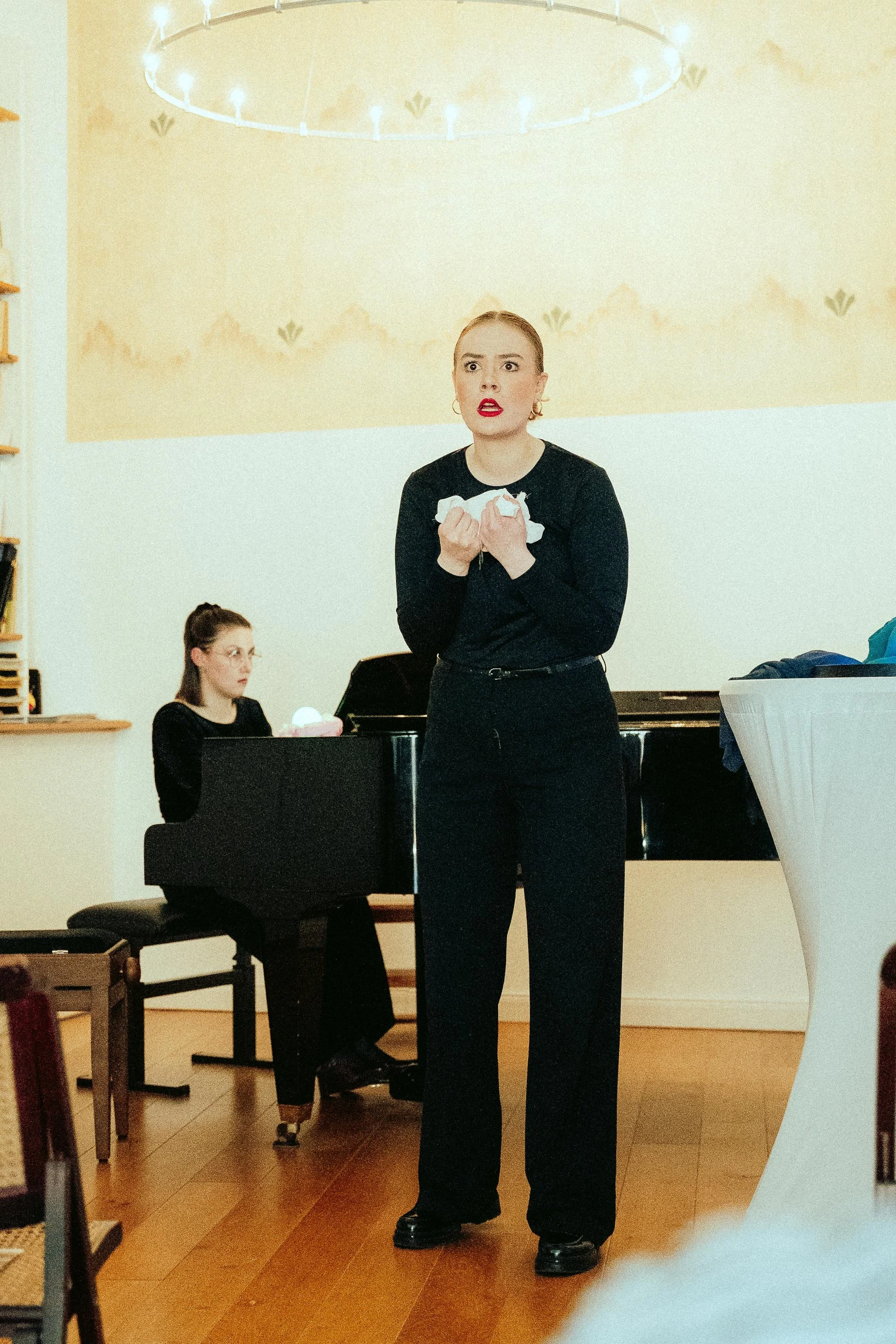 Eine Frau singt in einem Raum mit einem Klavier im Hintergrund. Sie trägt schwarze Kleidung und rote Lippen. Im Hintergrund sitzt eine weitere Person am Klavier.