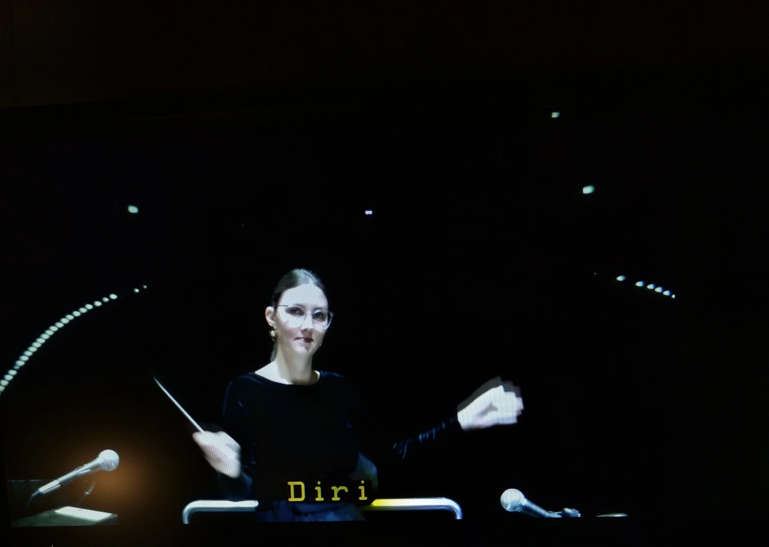 Eine Frau mit Brille und schwarzen Kleid am Pult, im Hintergrund Dunkelheit und Lichter, Untertitel zeigt den Namen 'Diri'.