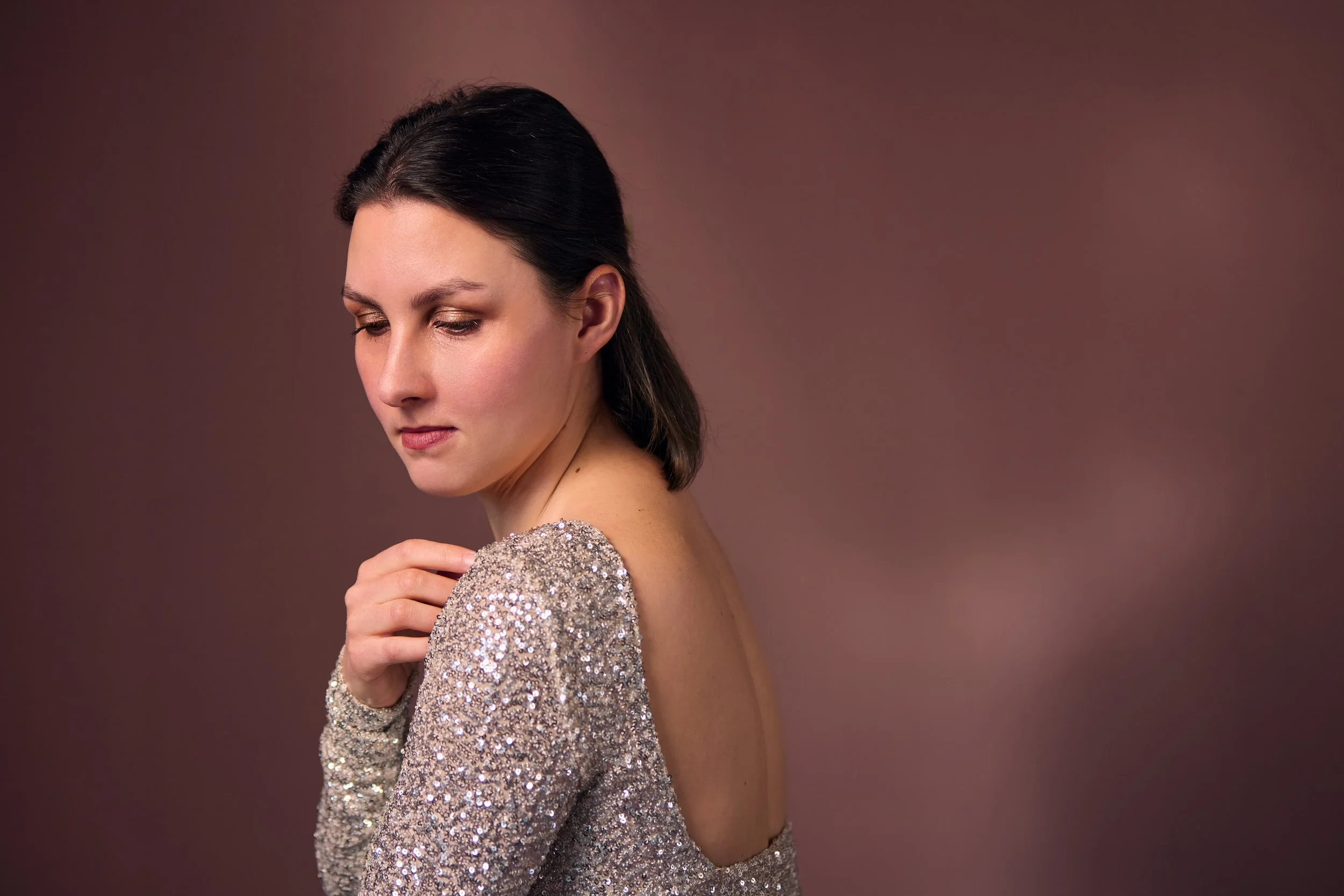 Ein Porträt einer Frau mit dunklem Haar, die eine glitzernde, silberne Abendrobe trägt. Sie blickt nach unten und hat eine nachdenkliche Haltung.