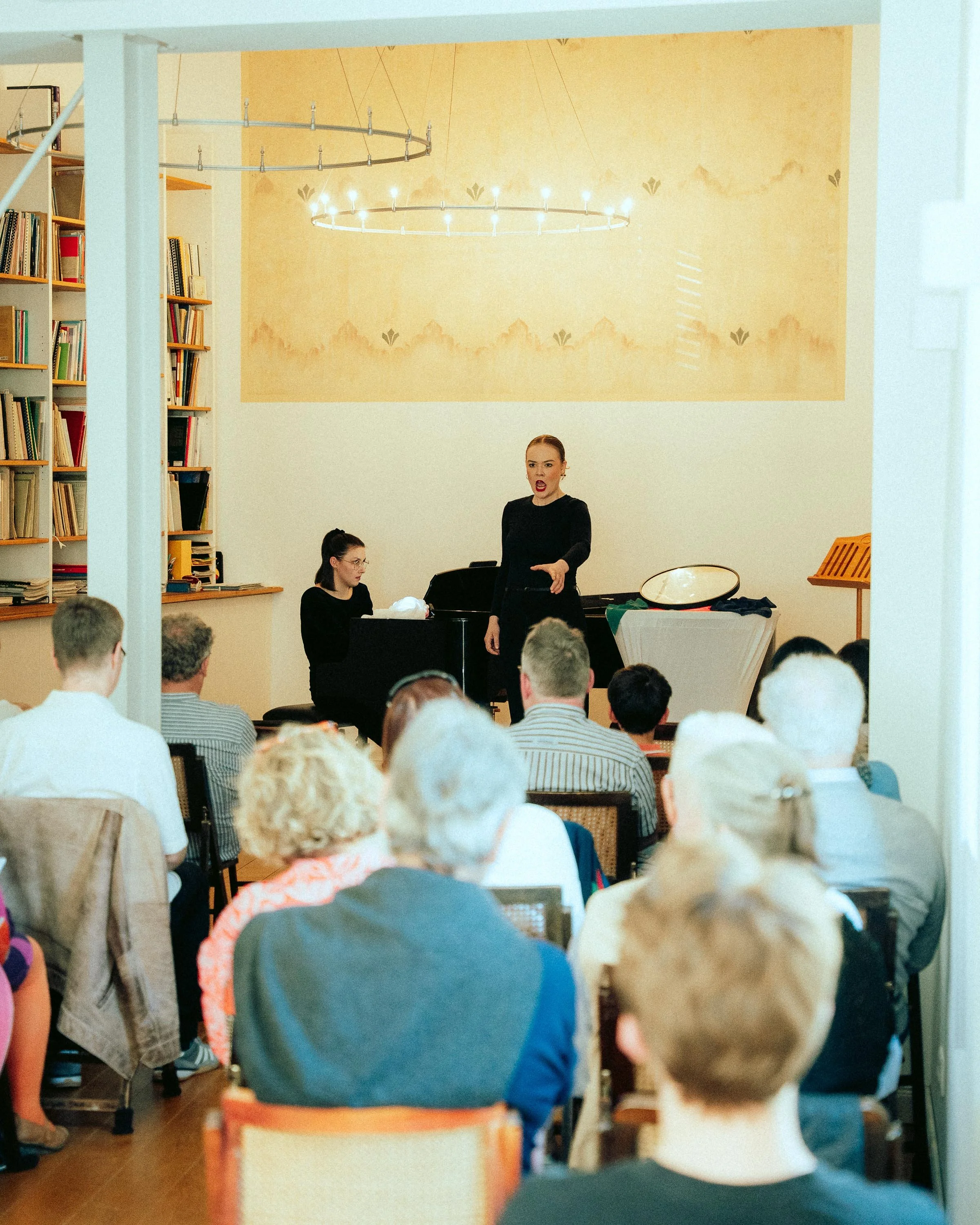 Ein Sprecher bei einem Konzert, umgeben von einem Publikum, in einem Raum mit Bücherregalen und einer kunstvollen Deckenleuchte.