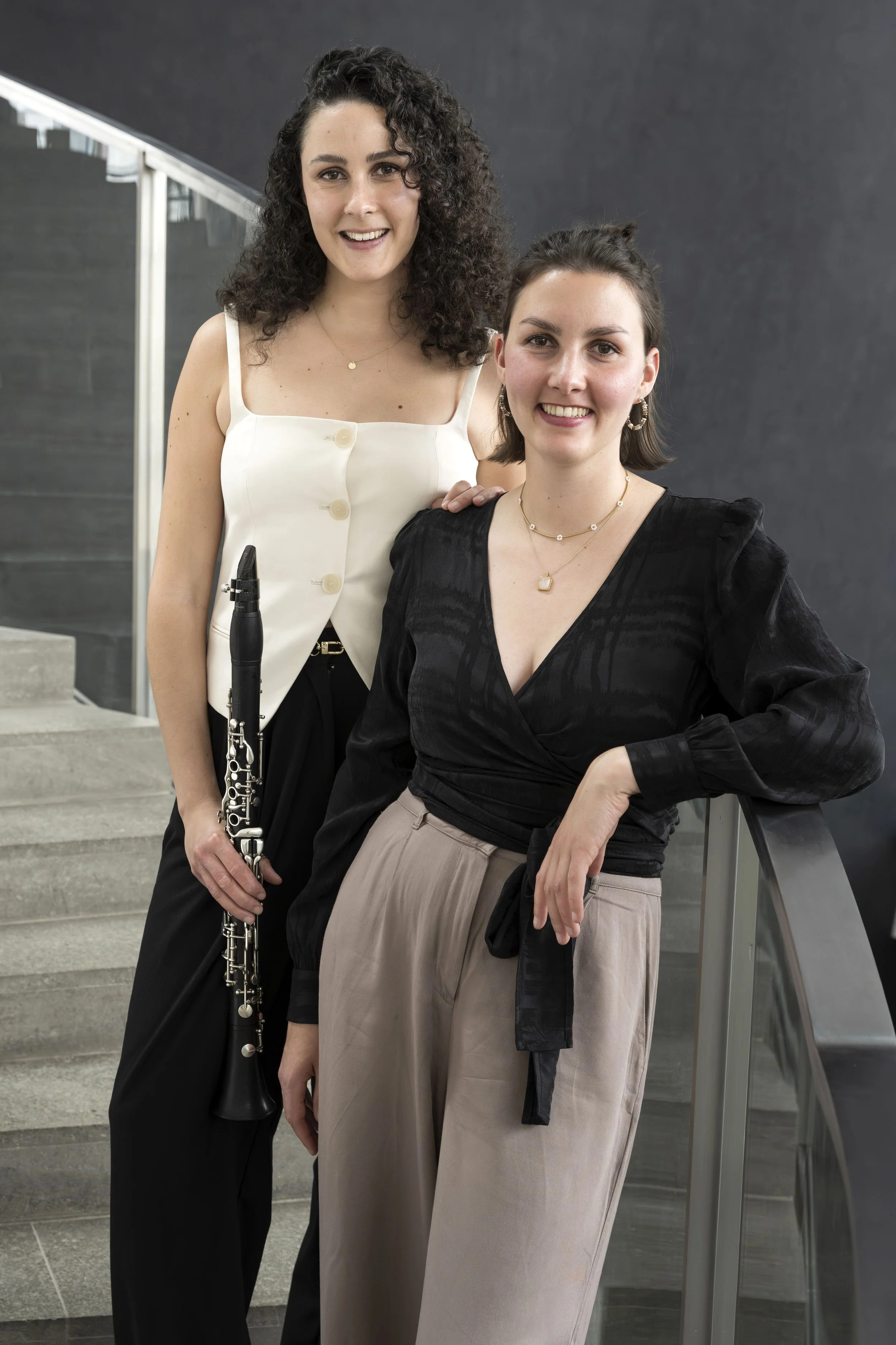 Zwei junge Frauen, eine mit dunkel, lockigem Haar, die andere mit braunen, glatten Haaren, posieren auf einer Treppe. Die eine hält eine Klarinette, beide lächeln und tragen elegante Kleidung.