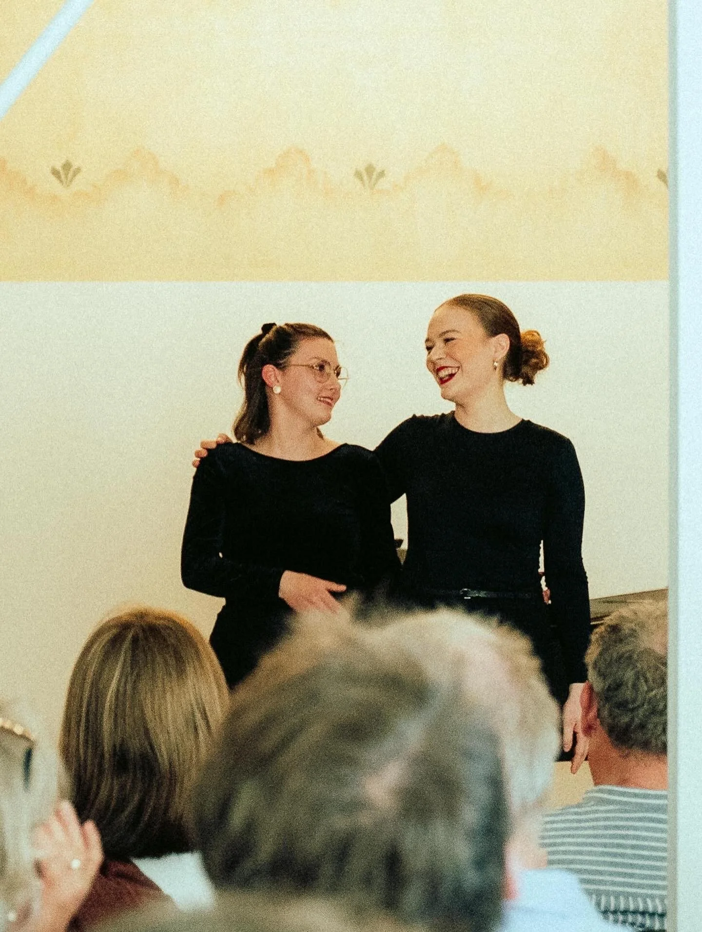 Zwei Frauen stehen auf einer Bühne, lachen und umarmen sich, während die Menschen im Publikum zuschauen und applaudieren.