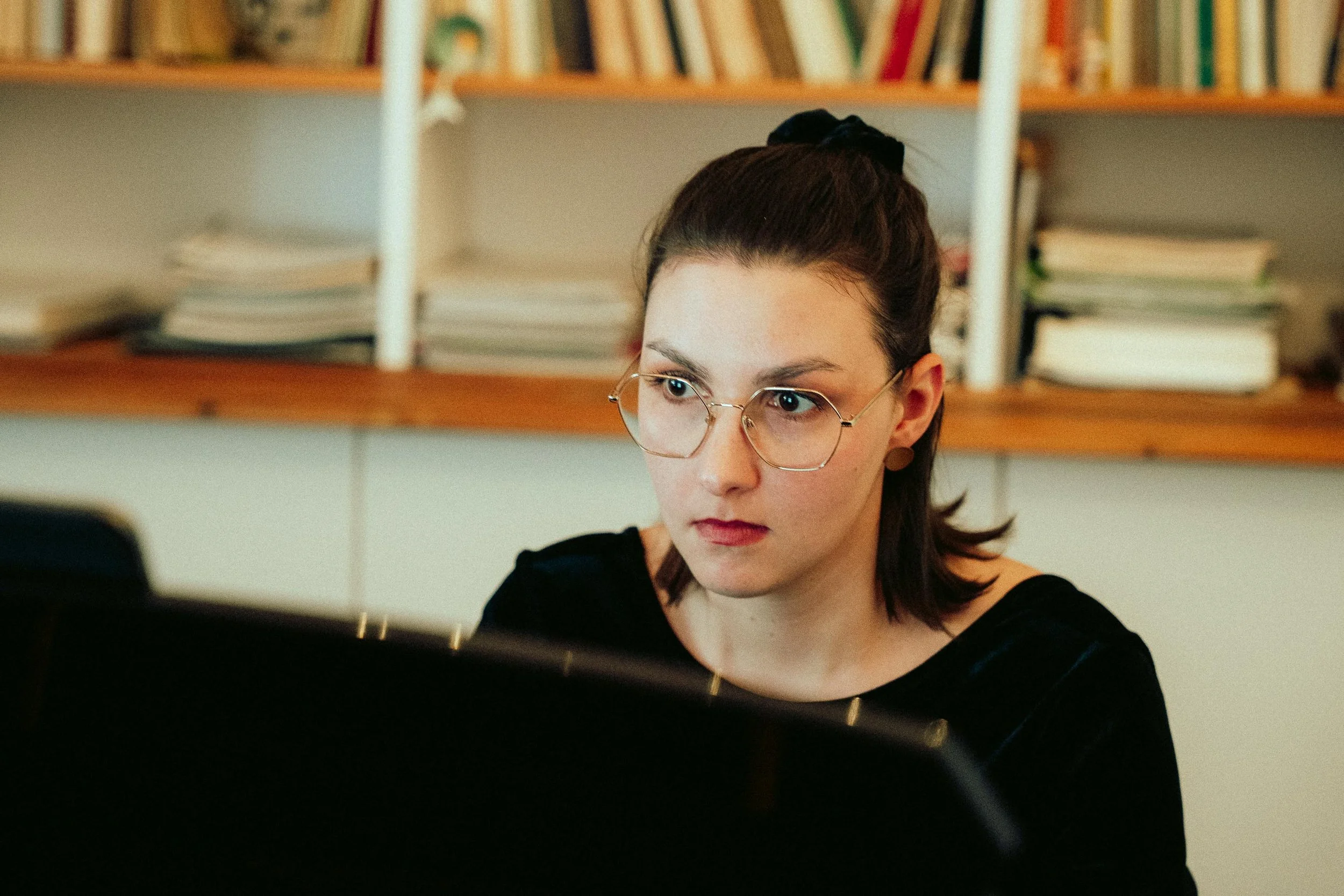 Eine junge Frau mit braunen Haaren, Brille und schwarzem Oberteil spielt Klavier in einem Raum mit Bücherregal im Hintergrund.