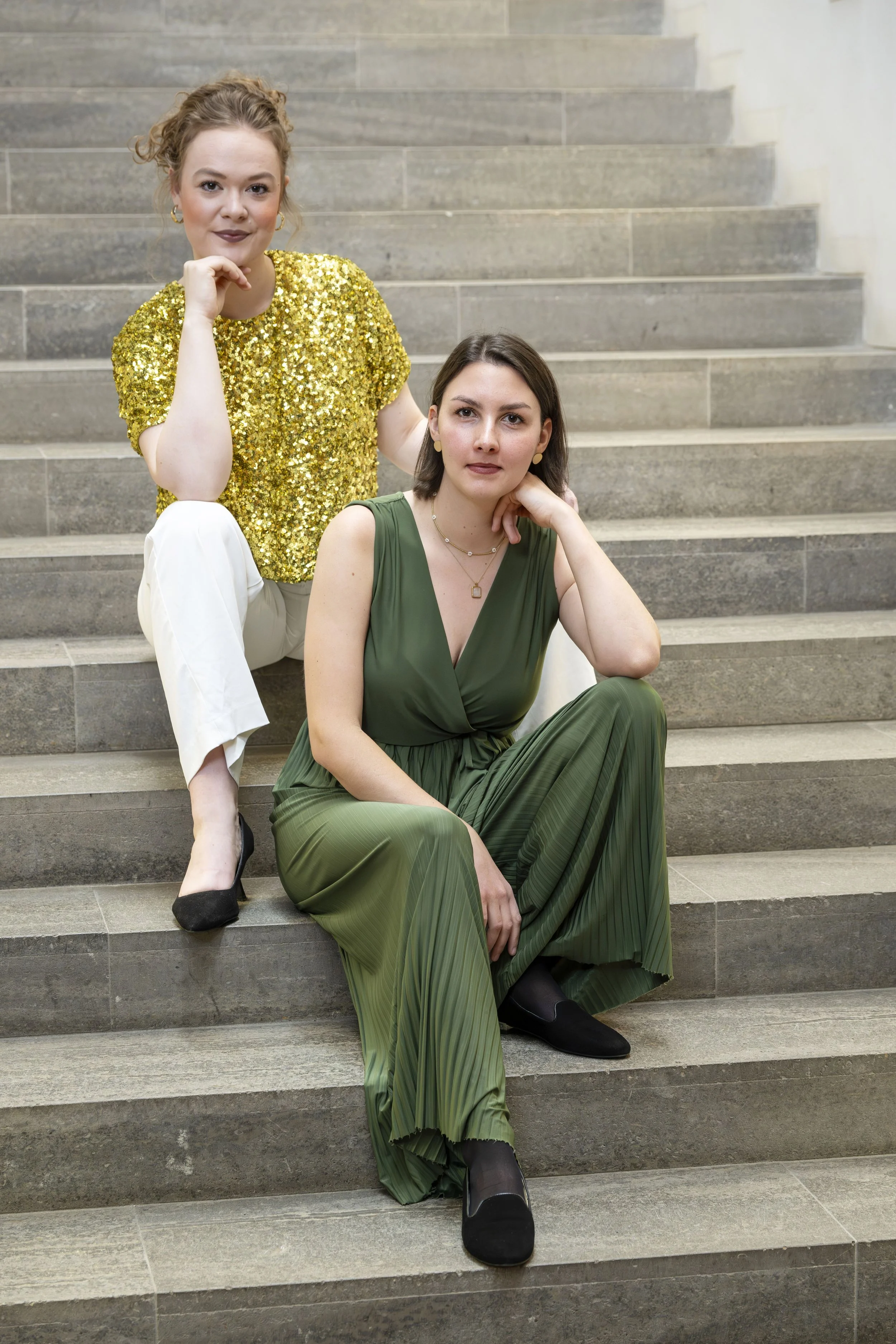 Zwei Frauen sitzen auf Steintreppen, die eine trägt ein grünes Kleid und die andere ein goldenes Paillettenkleid.