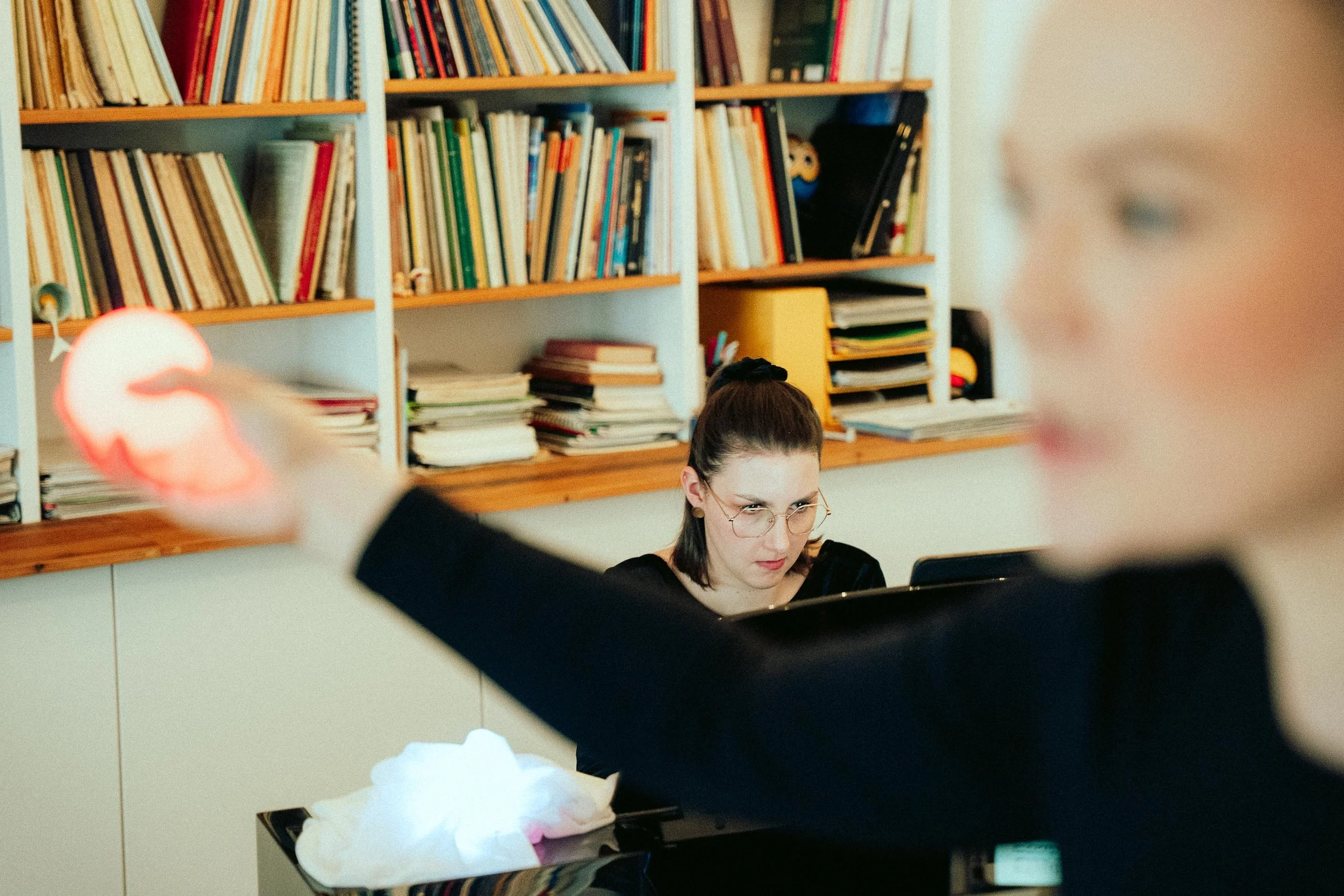Junge Frau spielt Klavier, im Hintergrund Bücherregale voller Bücher, andere Personen im Raum, Beleuchtung warm.