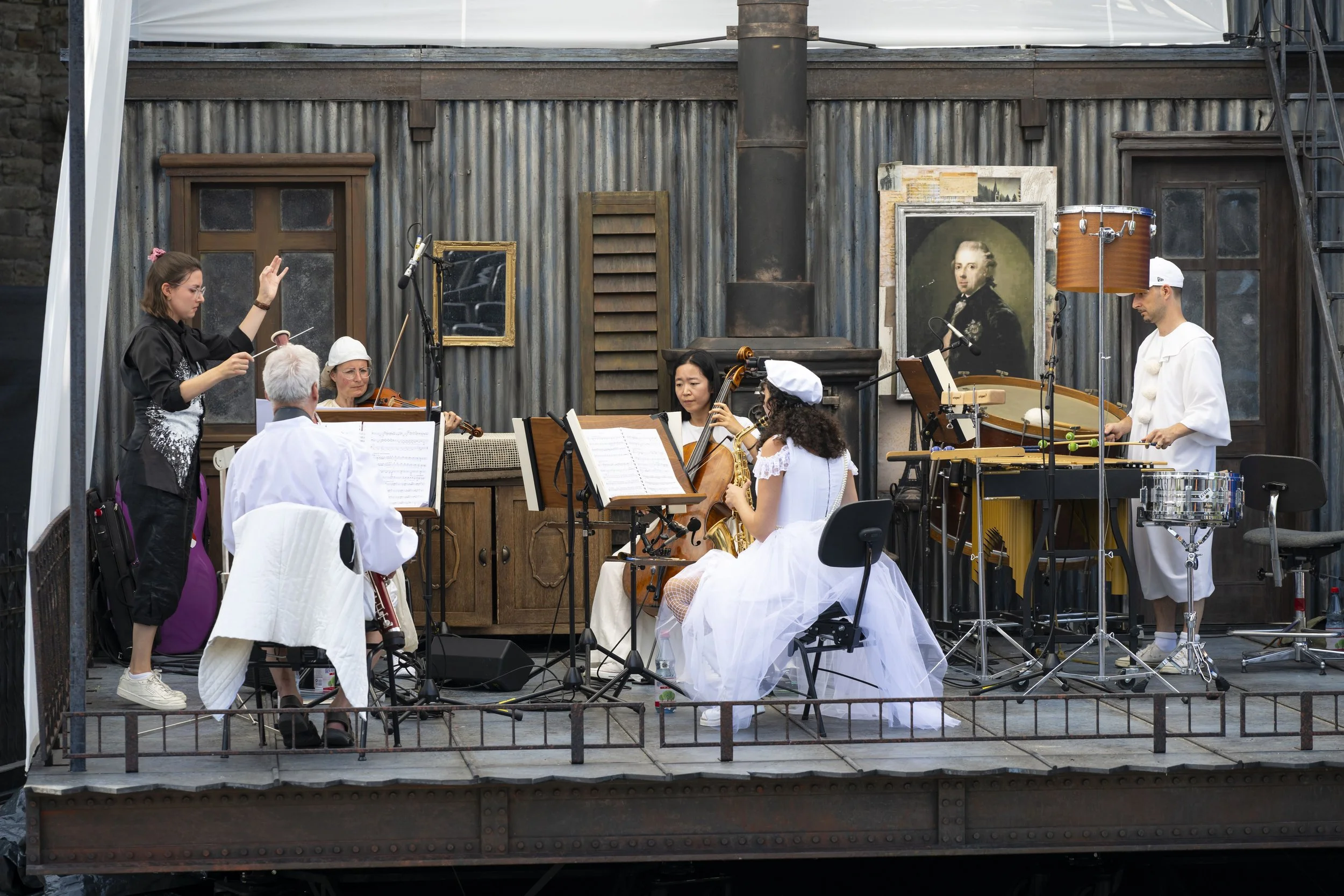 Ein Orchester probt auf einer Bühne im Freien, mit Musikern, Dirigentin und Instrumenten wie Geige, Cello, Schlagzeug und Percussion vor einem rustikalen Hintergrund mit alten Rahmen und Profilbildern.