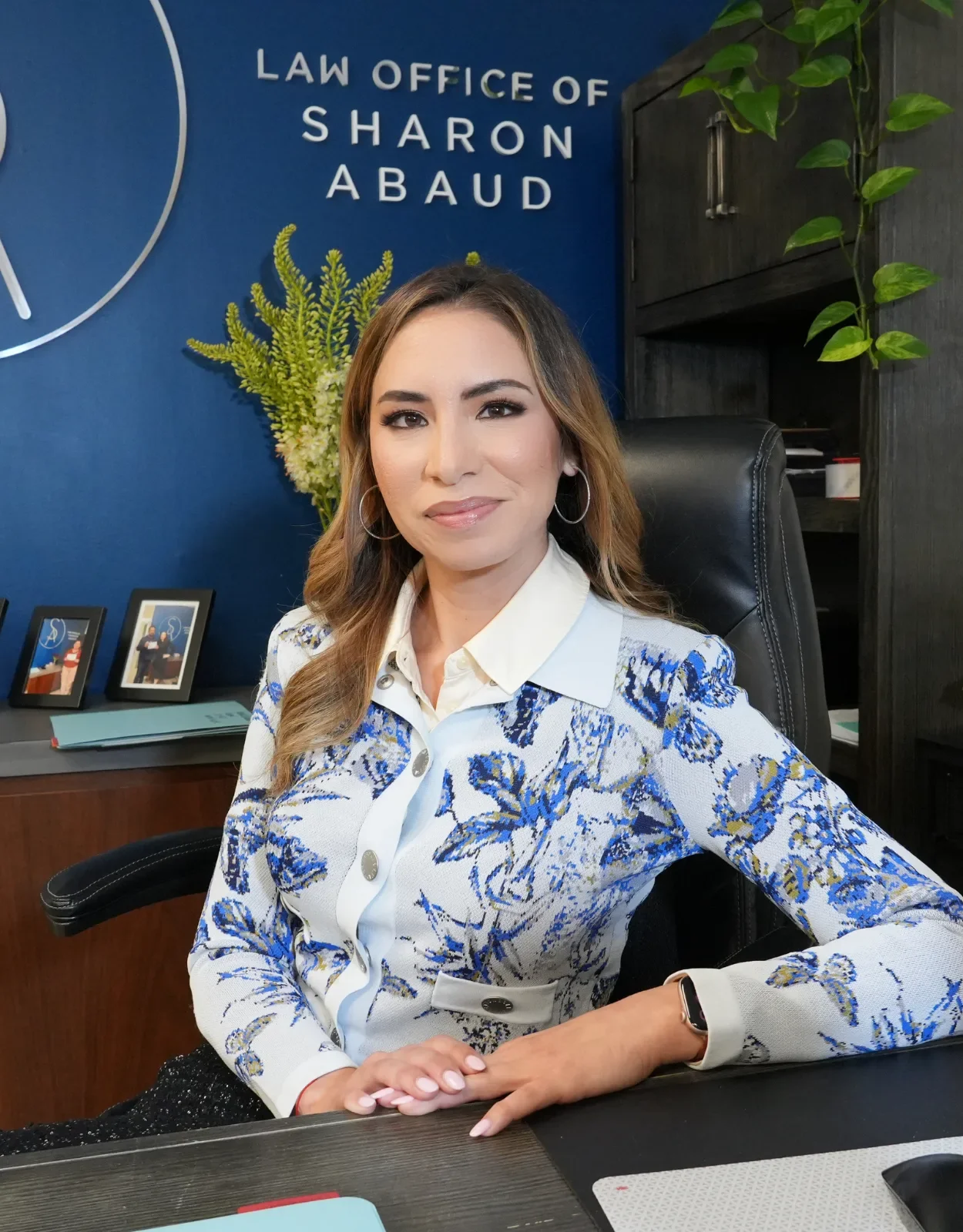 Mujer en una oficina con fondo azul que tiene un cartel que dice 'Law Office of Sharon Abaud', sentada en una silla con blusa blanca y estampada en azul y dorado, mirando a la cámara.