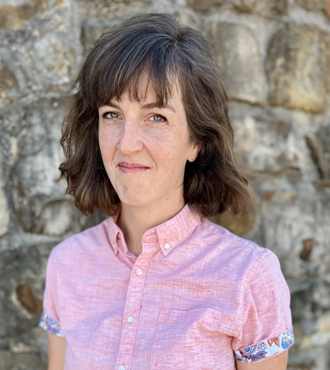 Portrait of a woman with shoulder-length brown hair, blue eyes, wearing a pink button-up shirt with patterned sleeve cuffs, standing outdoors in front of a stone wall.