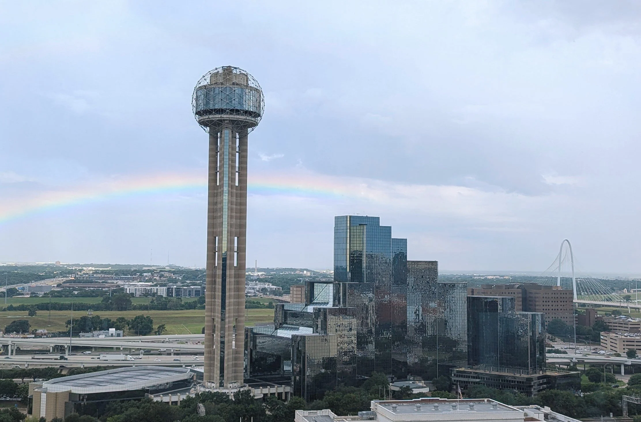 Dallas skyline with a tall observation tower featuring a spherical top, partially obscured by clouds, a rainbow in the background, and modern glass buildings, highway, and bridge in the foreground.