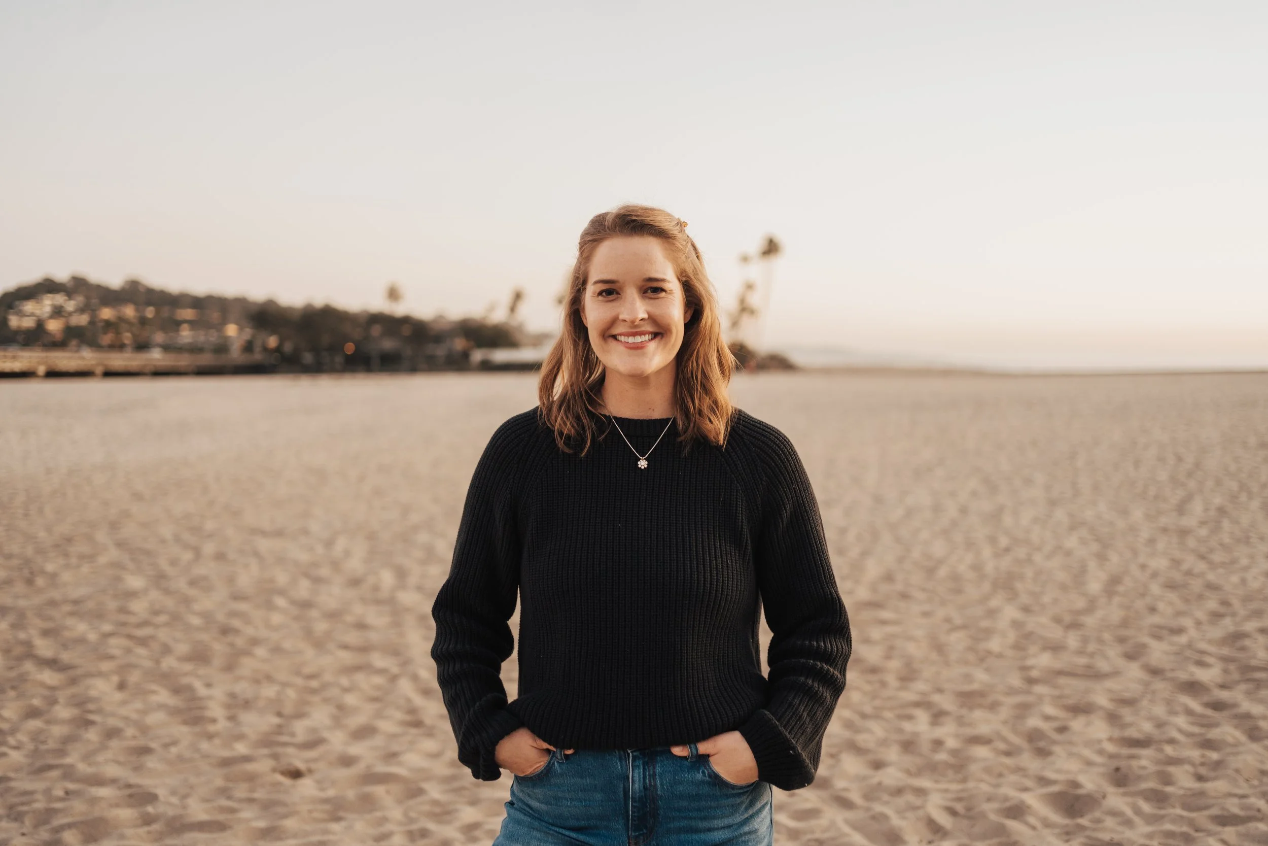 A smiling woman with brown hair standing on a sandy beach during sunset, wearing a black sweater and jeans, with a cloudy sky and distant shoreline in the background.