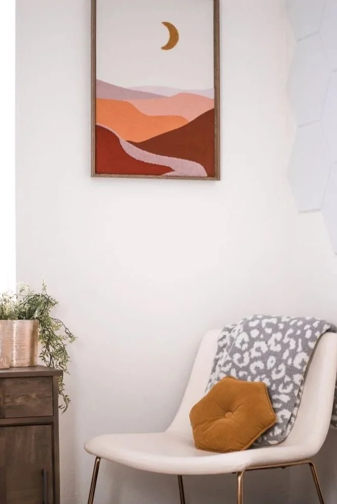 Interior of a cozy living space with a white chair, a mustard yellow pillow, a gray and white patterned throw blanket, a wooden side table with a potted plant, and a framed abstract landscape painting with a crescent moon hanging above.