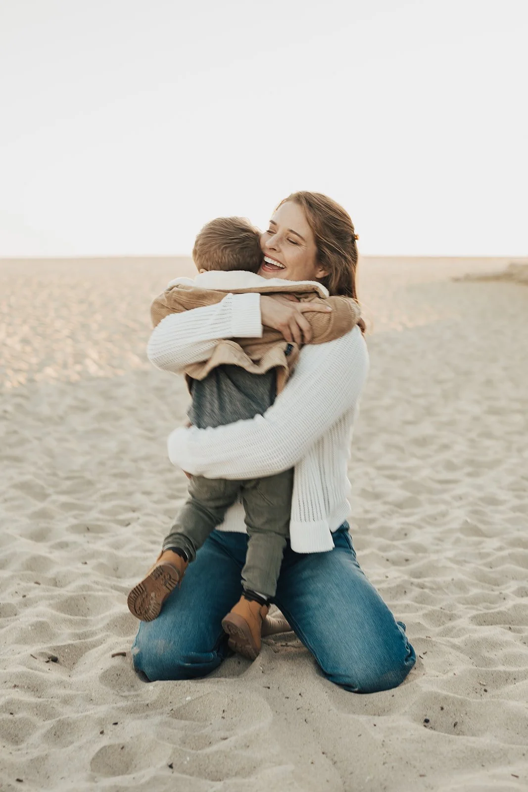 A woman kneeling on sandy beach, hugging a young boy, both smiling, with clear sky and beach in background.