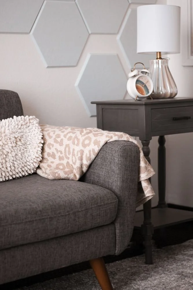 A living room corner with a gray fabric sofa, a cream and beige animal print throw blanket, a textured white pillow, and a dark gray side table with a silver lamp and white clock.
