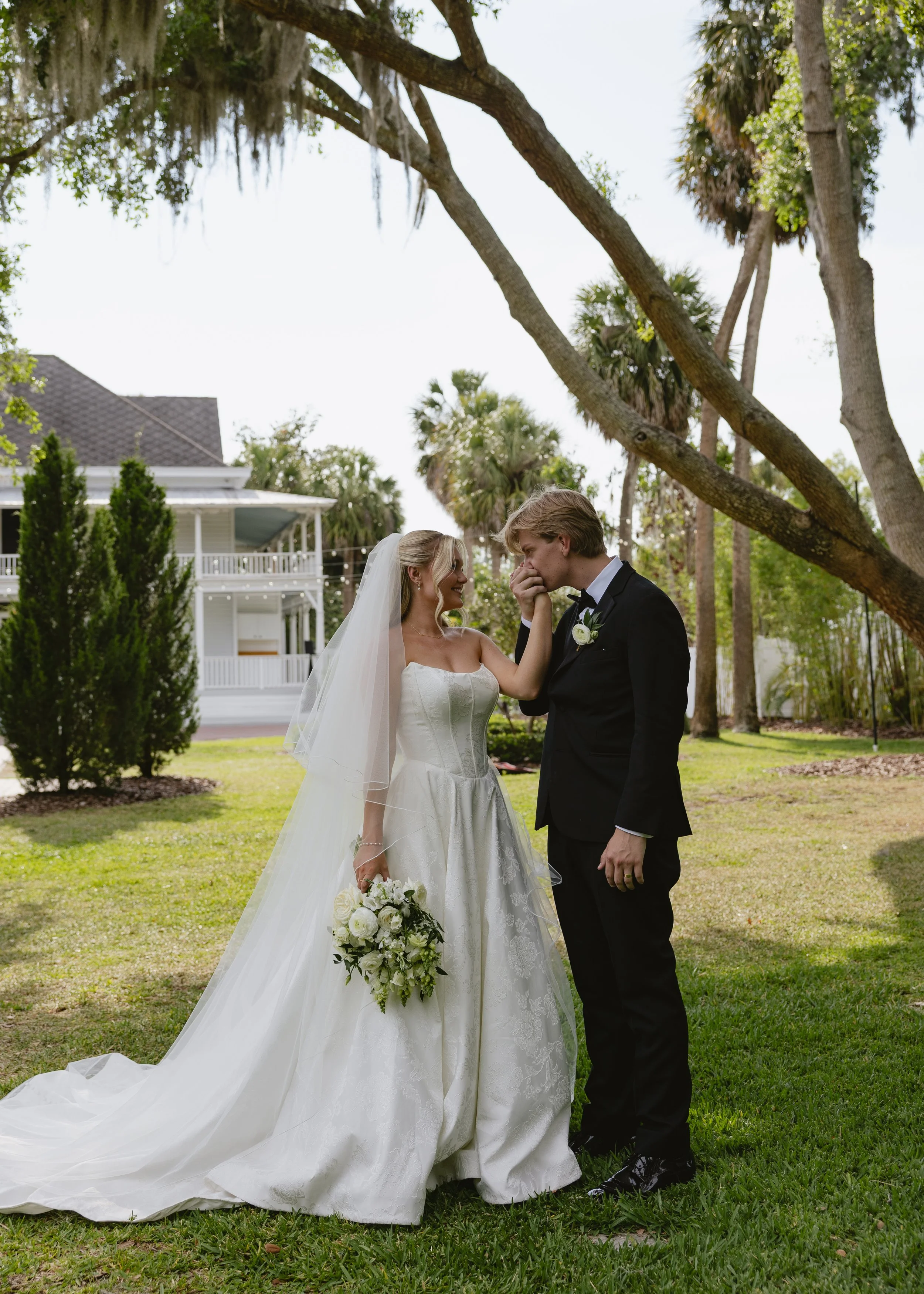 A bride and groom standing close together outdoors, with the groom touching the bride's face, during their wedding ceremony on a grassy area with trees and a white house in the background.
