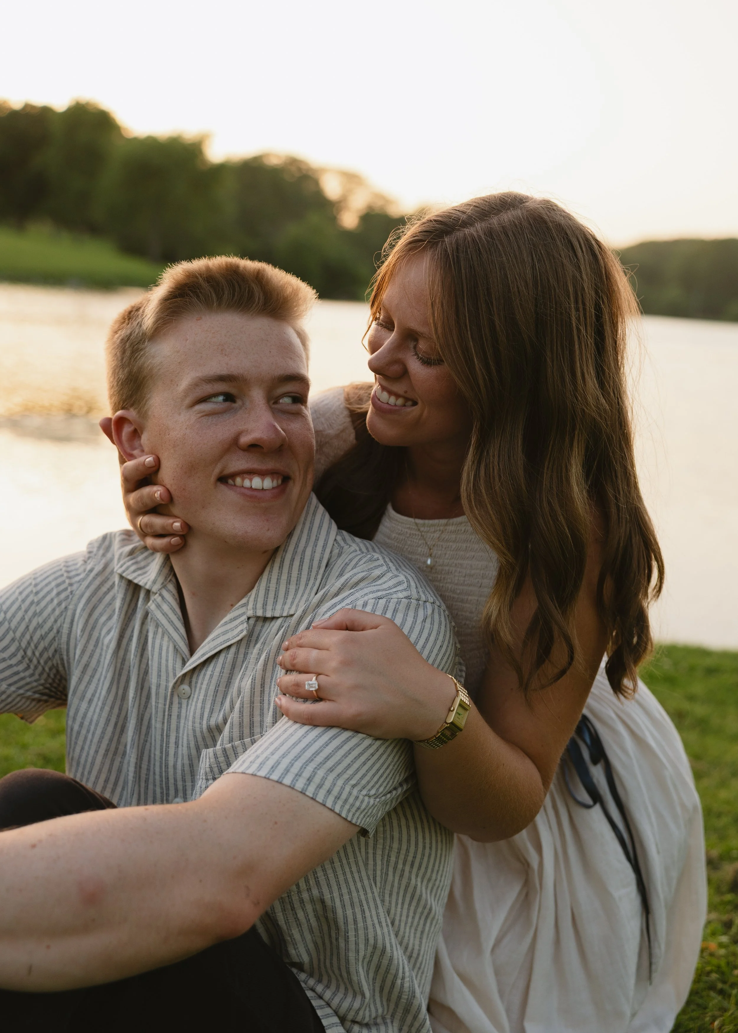 A woman and a teenage boy sitting outdoors near a body of water, smiling and enjoying each other's company during sunset.