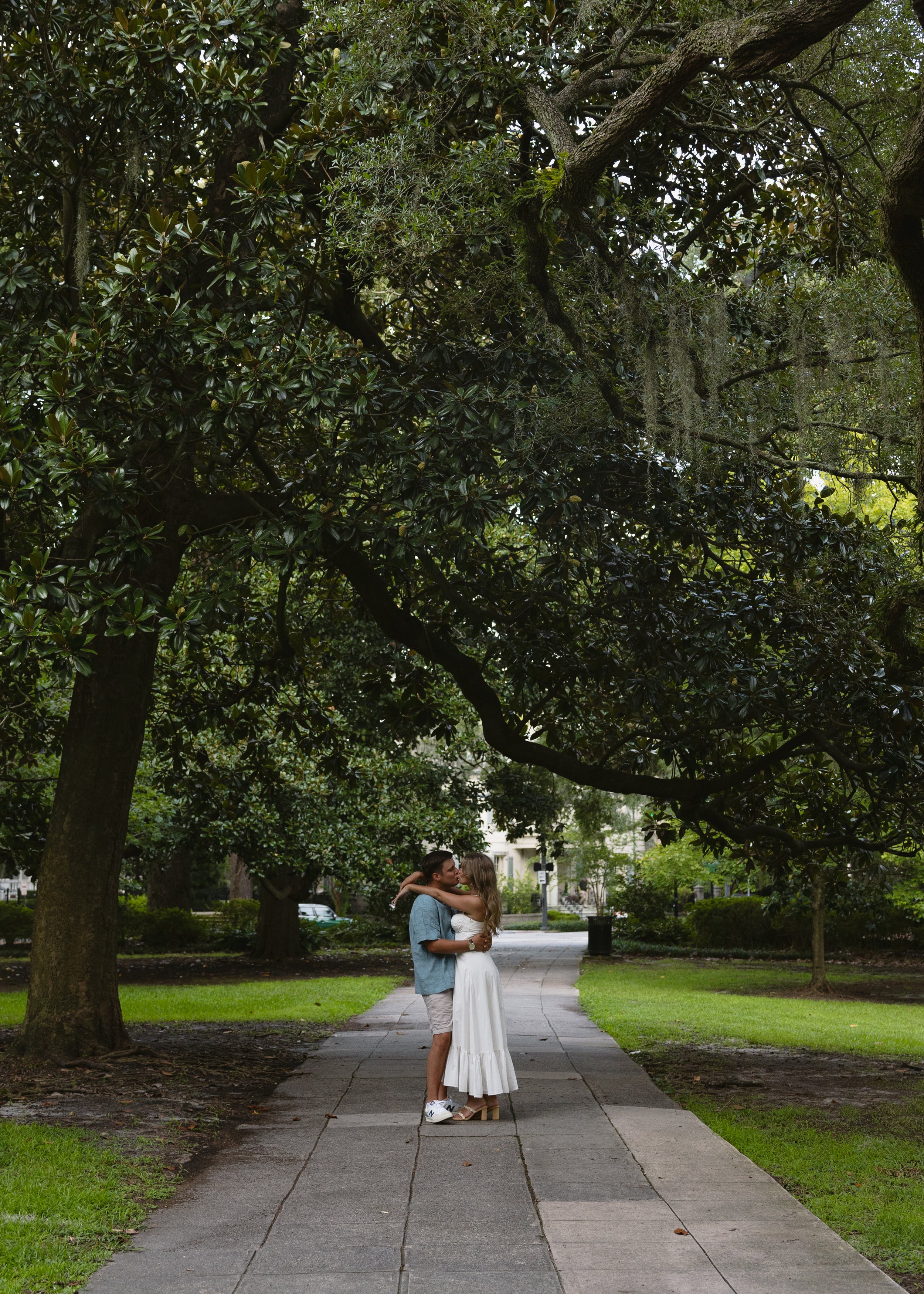 A couple kissing on a sidewalk in a park with lush green trees and grass.