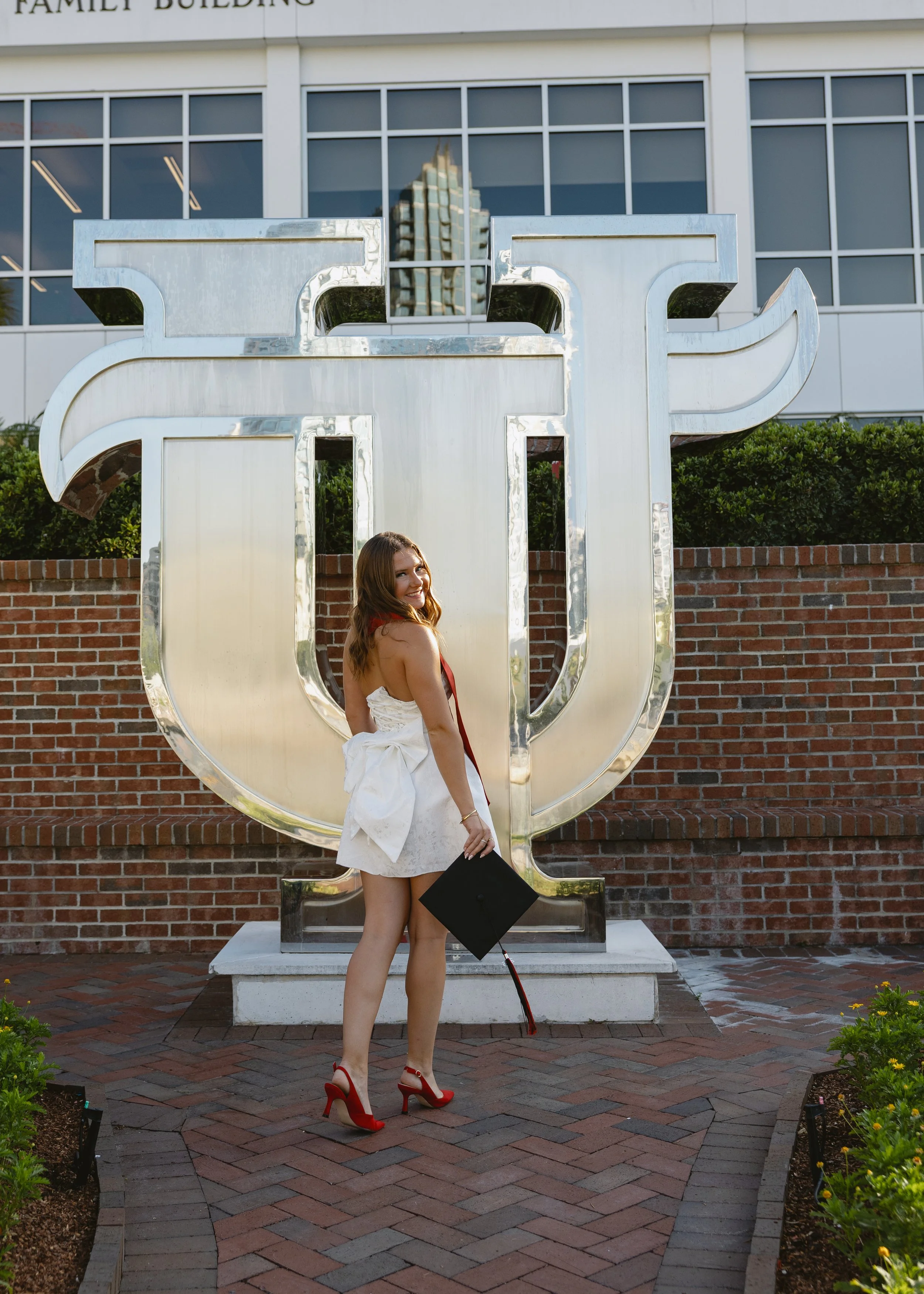 A young woman in a white dress with a large white bow and red high-heeled shoes stands holding a diploma, smiling, in front of a large metallic shield-like sculpture, with a brick building and greenery behind her.