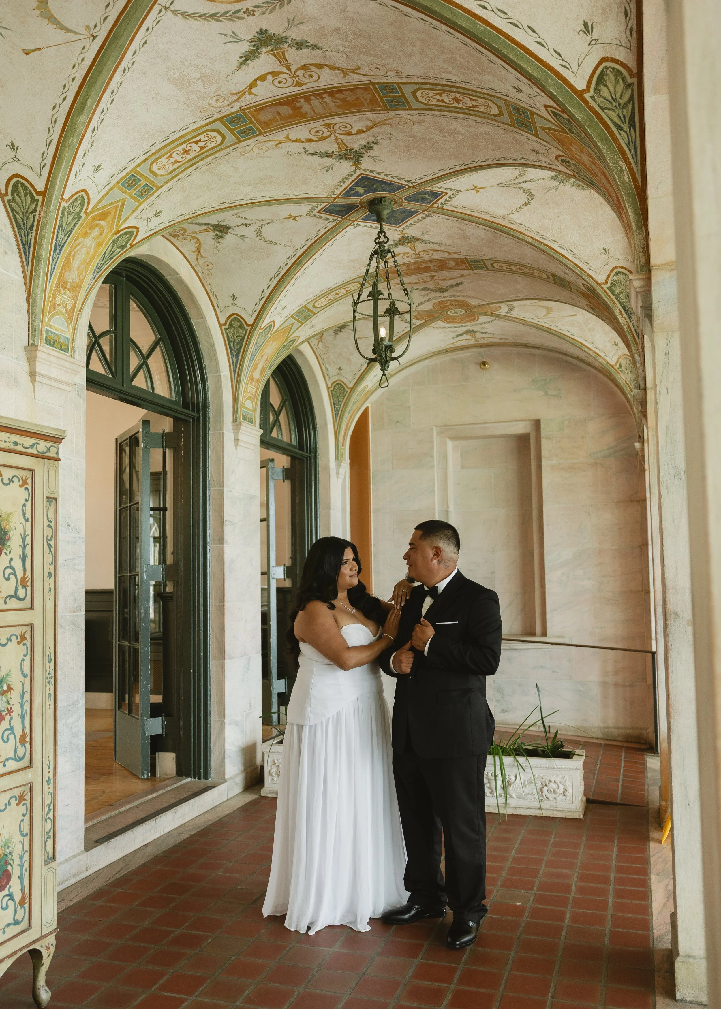 A newlywed couple dressed in wedding attire standing in an elegant, historic corridor with painted vaulted ceilings and arched windows, sharing a moment together.