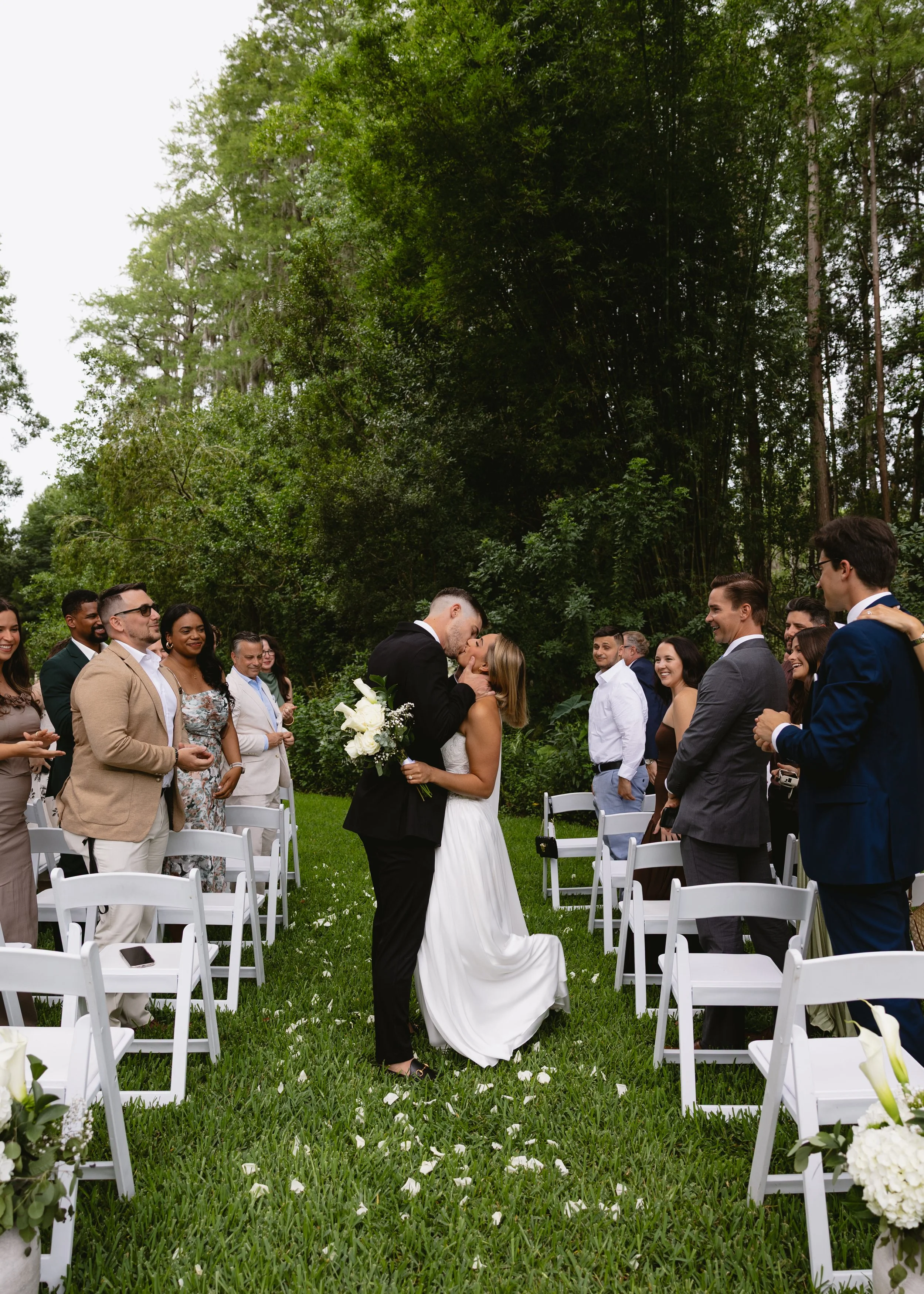 A bride and groom kissing during their outdoor wedding ceremony, surrounded by friends and family under a large green tree.