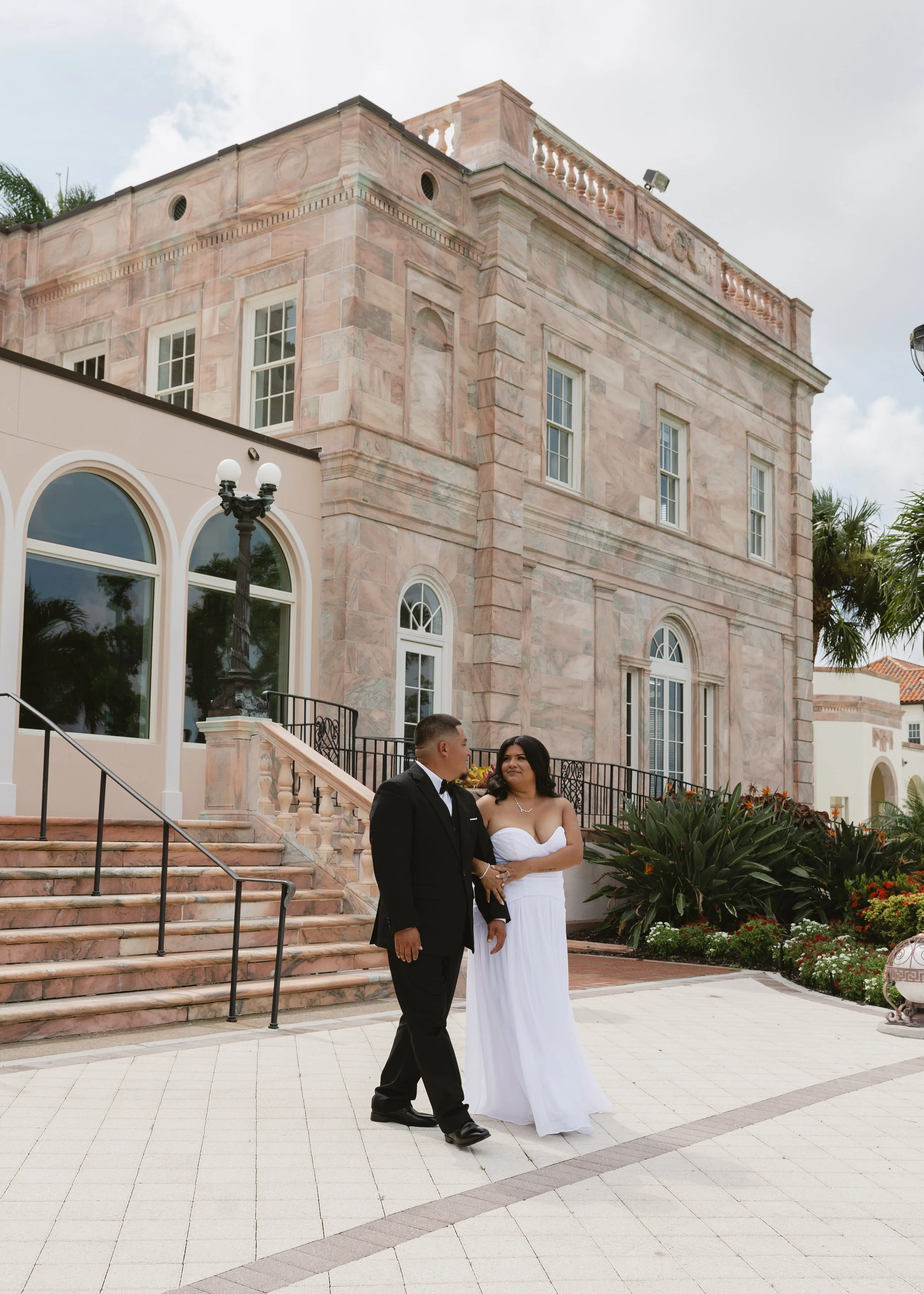 A couple in wedding attire standing outside in front of a pink stone building with large arched windows and a staircase. The man is dressed in a black tuxedo, and the woman is wearing a strapless white wedding gown. There are plants and flowers nearb