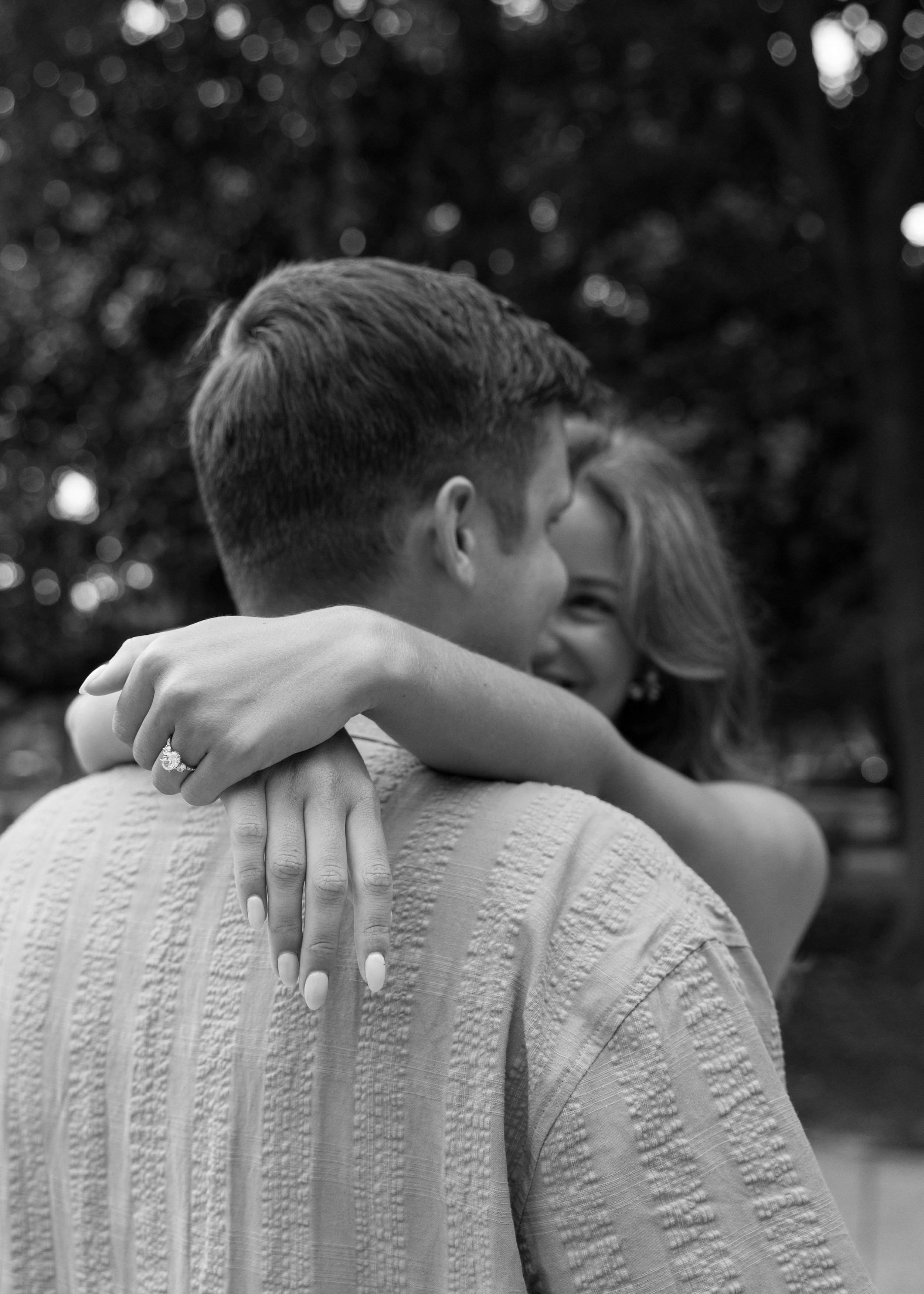 A black-and-white photo of a couple embracing, focusing on the woman's hand with a ring and their faces pressed close together.