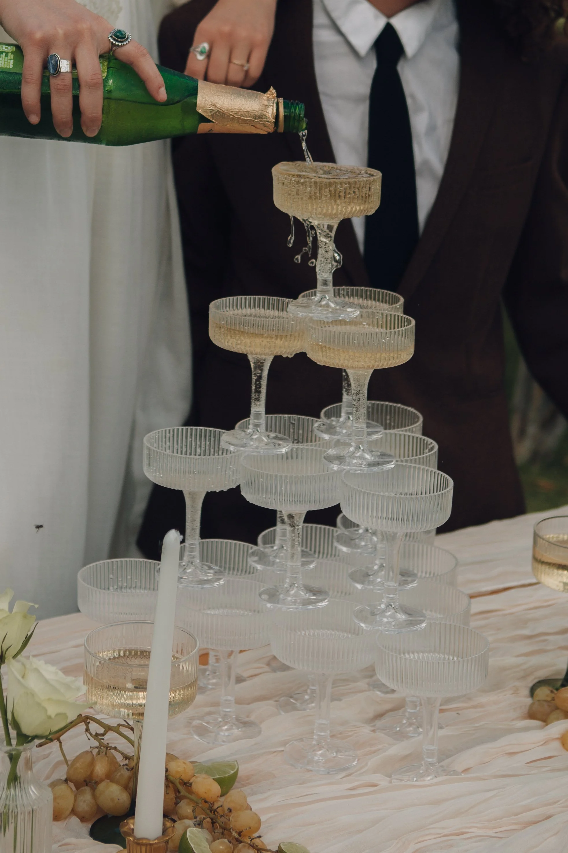 Chandeliers arranged in a tower with champagne bottles pouring into glasses, a partially visible couple in formal attire, and a table decorated with flowers and grapes at an event or celebration.
