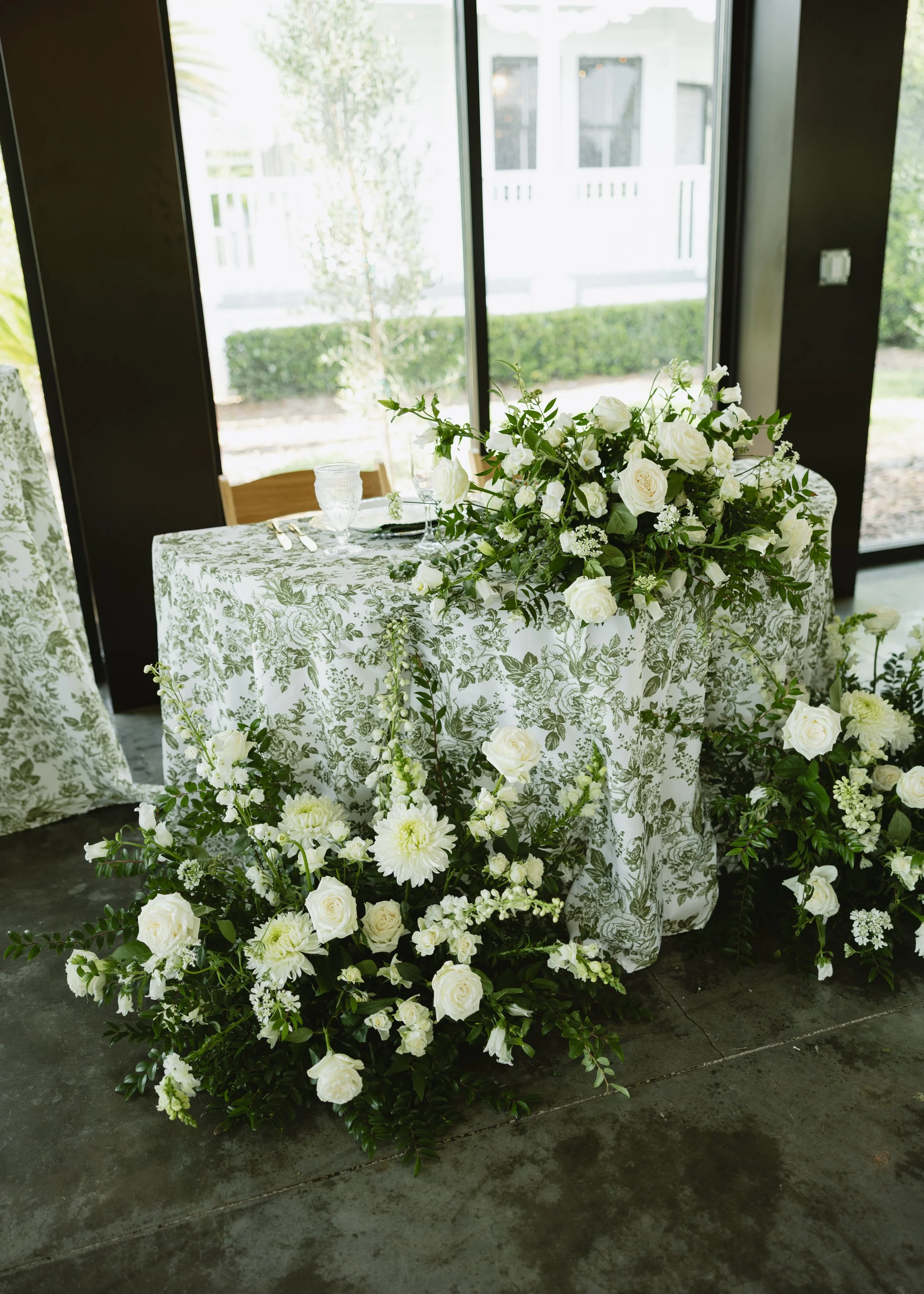 A table with a floral patterned tablecloth, decorated with a large bouquet of white flowers, set in front of large glass doors with a view of a garden outside.
