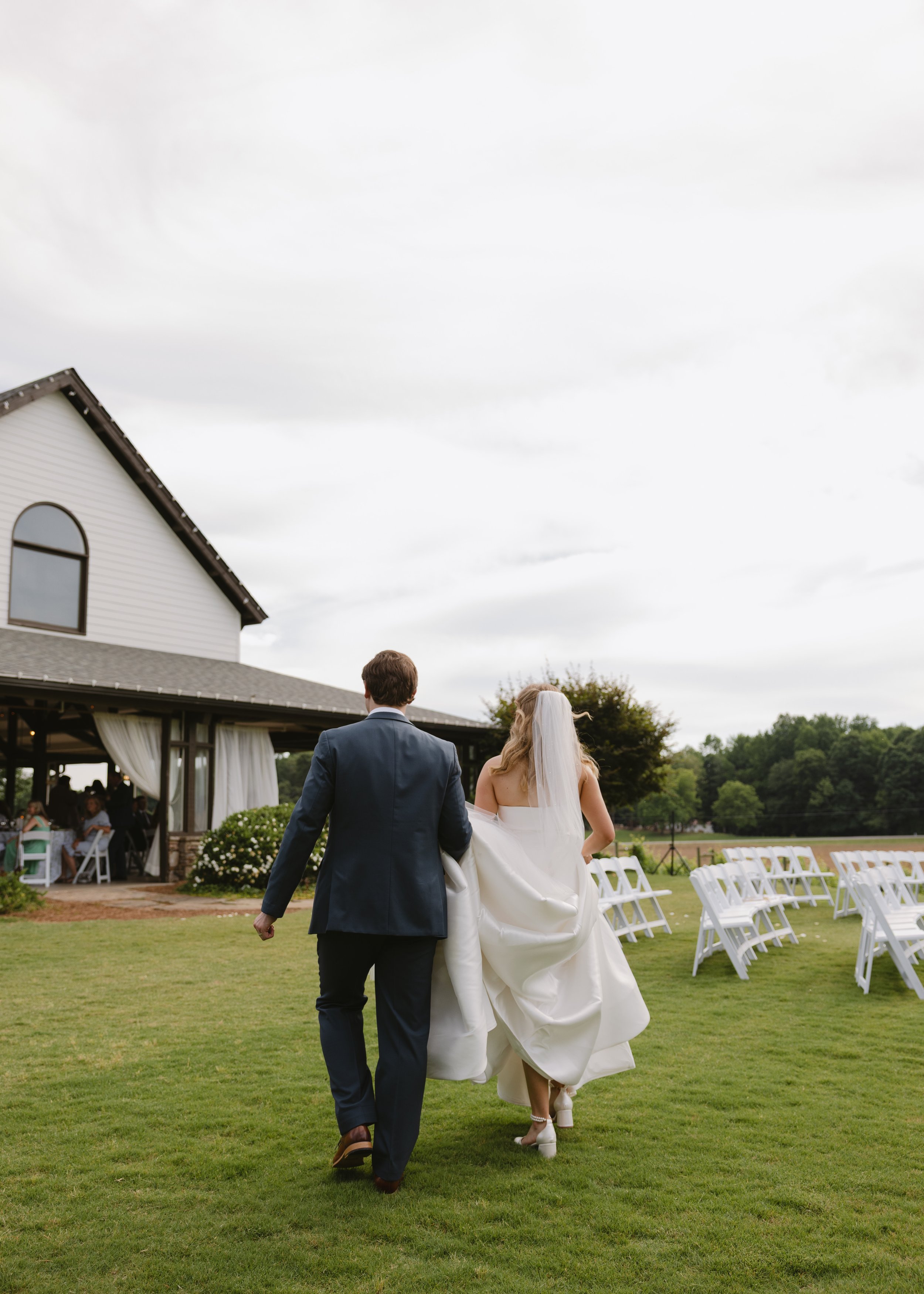 A bride and groom walking on a grassy area during their outdoor wedding ceremony, with chairs and a white building with a large window in the background.