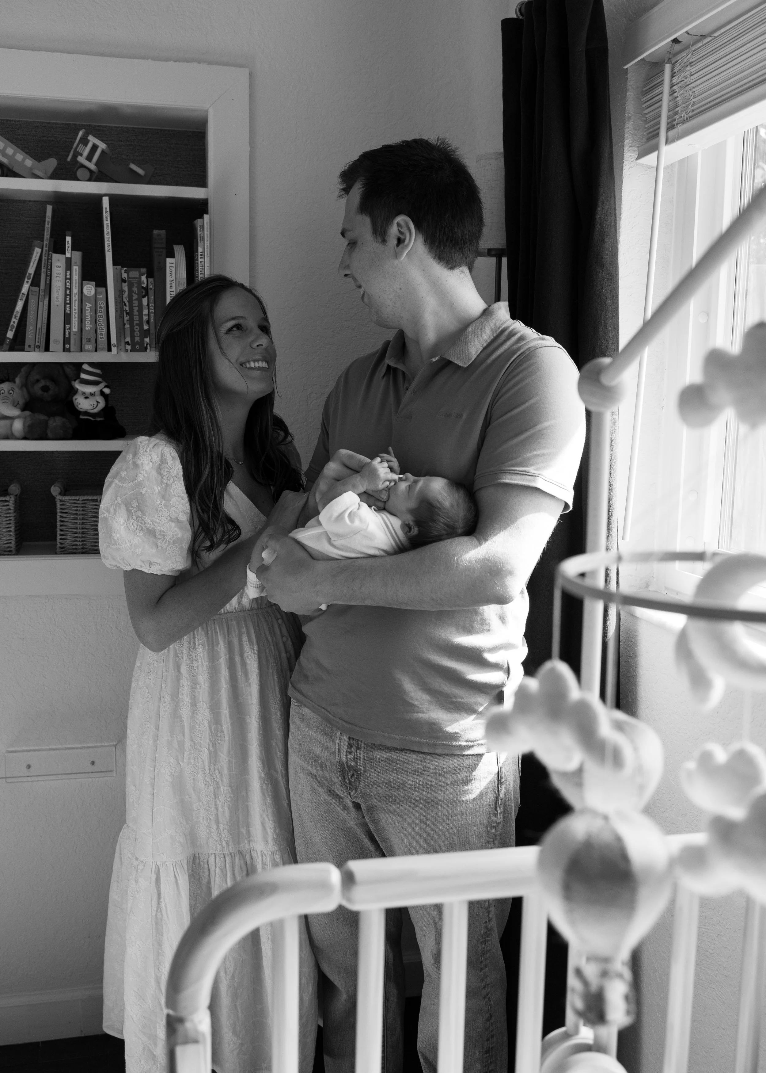A family in a hospital room. A woman dressed in white, holding a newborn baby, looks up at a man who is smiling at her. The man is standing in front of a window, near a curtain and medical equipment.