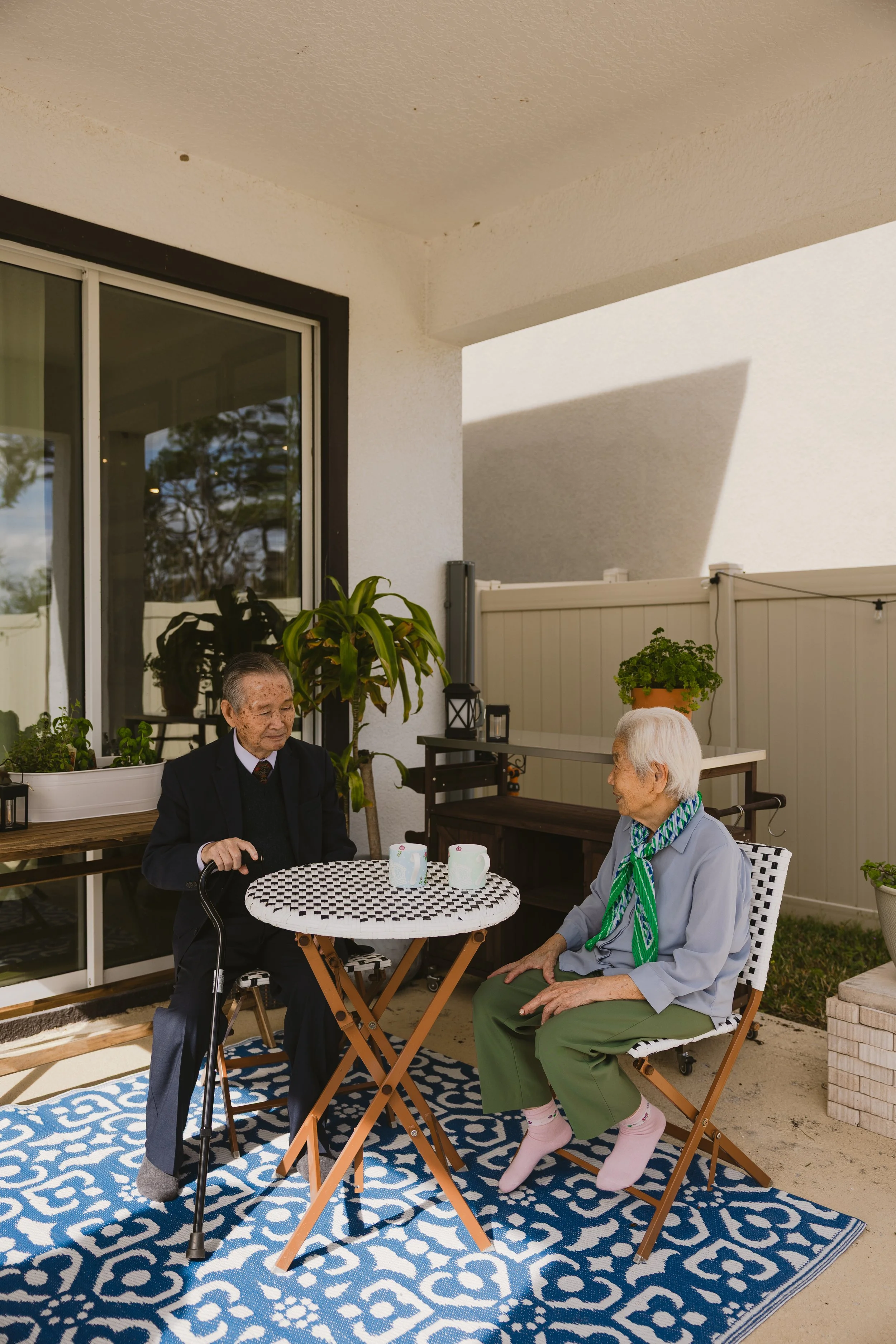 Two elderly women sitting at a small outdoor table on a patio, engaged in conversation. One woman has a cane and is wearing a blazer, while the other is dressed in a gray shirt, green pants, and a green scarf. The patio has potted plants, a patterned