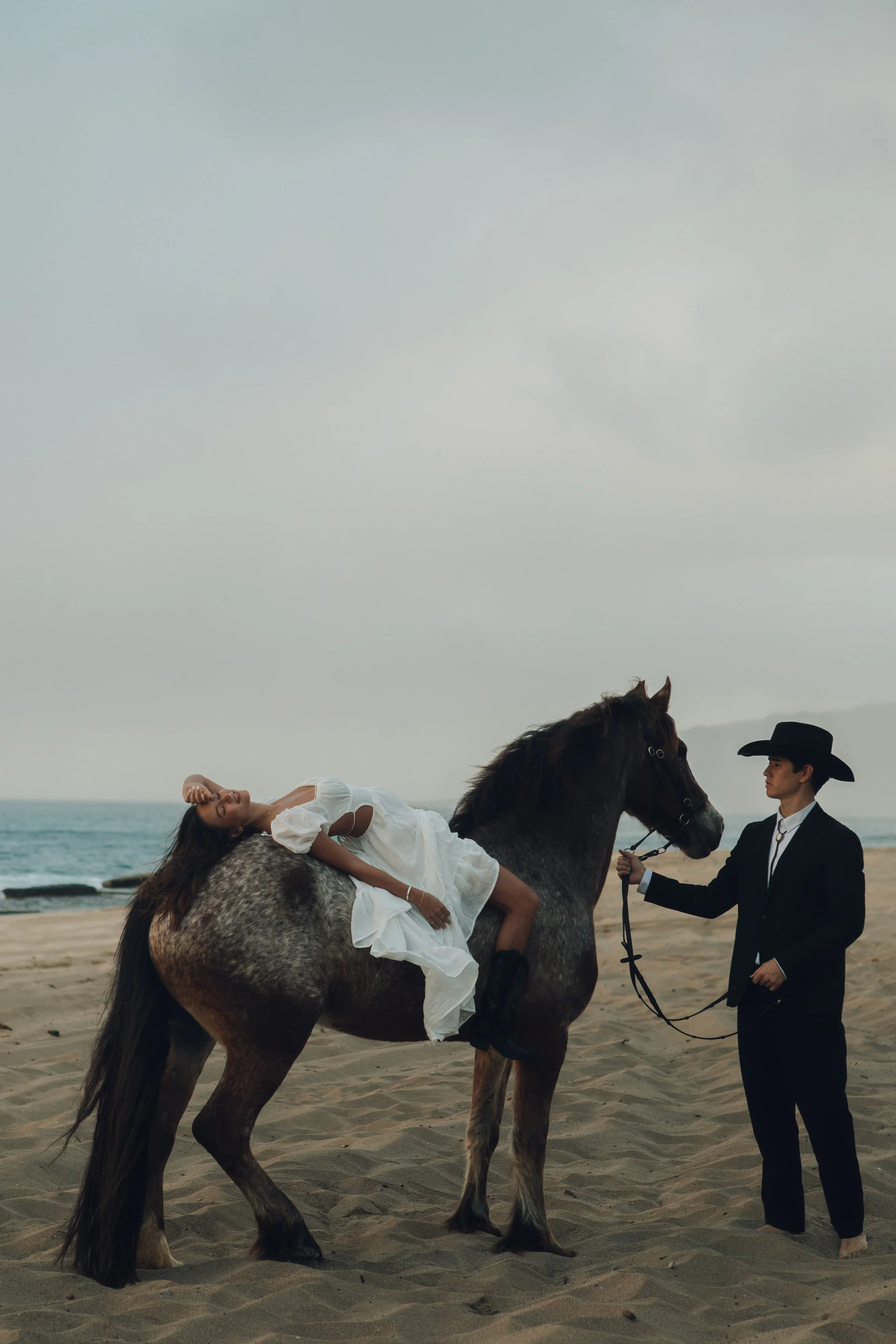 A woman in a white dress lying on a brown horse on a sandy beach, with a man in a black suit and hat holding the horse's reins.