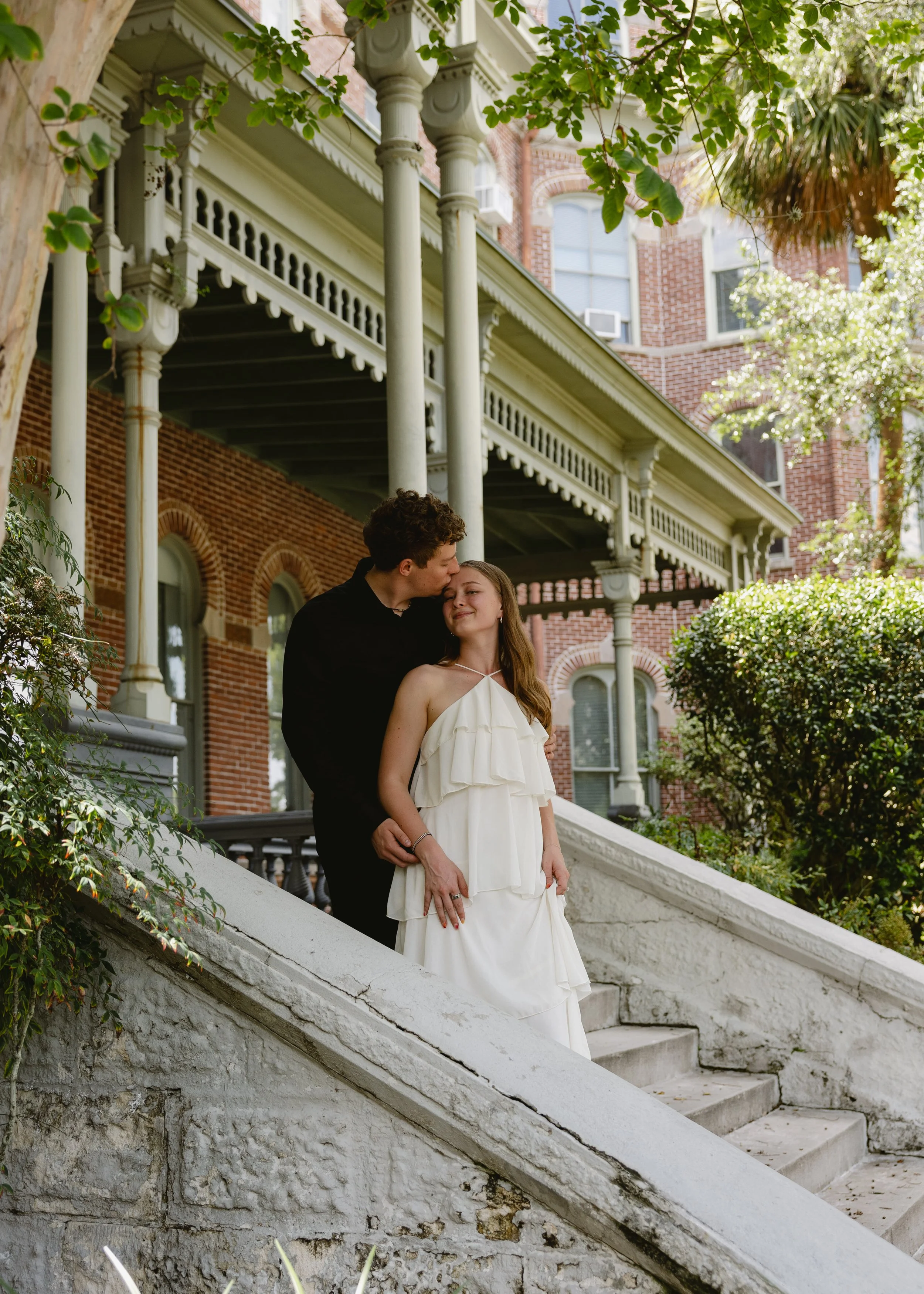 A young couple standing close together on the steps of a historic brick building with a large porch, surrounded by greenery. The man is kissing the woman on the forehead, and she is smiling with her eyes closed. The woman is wearing a white dress, an