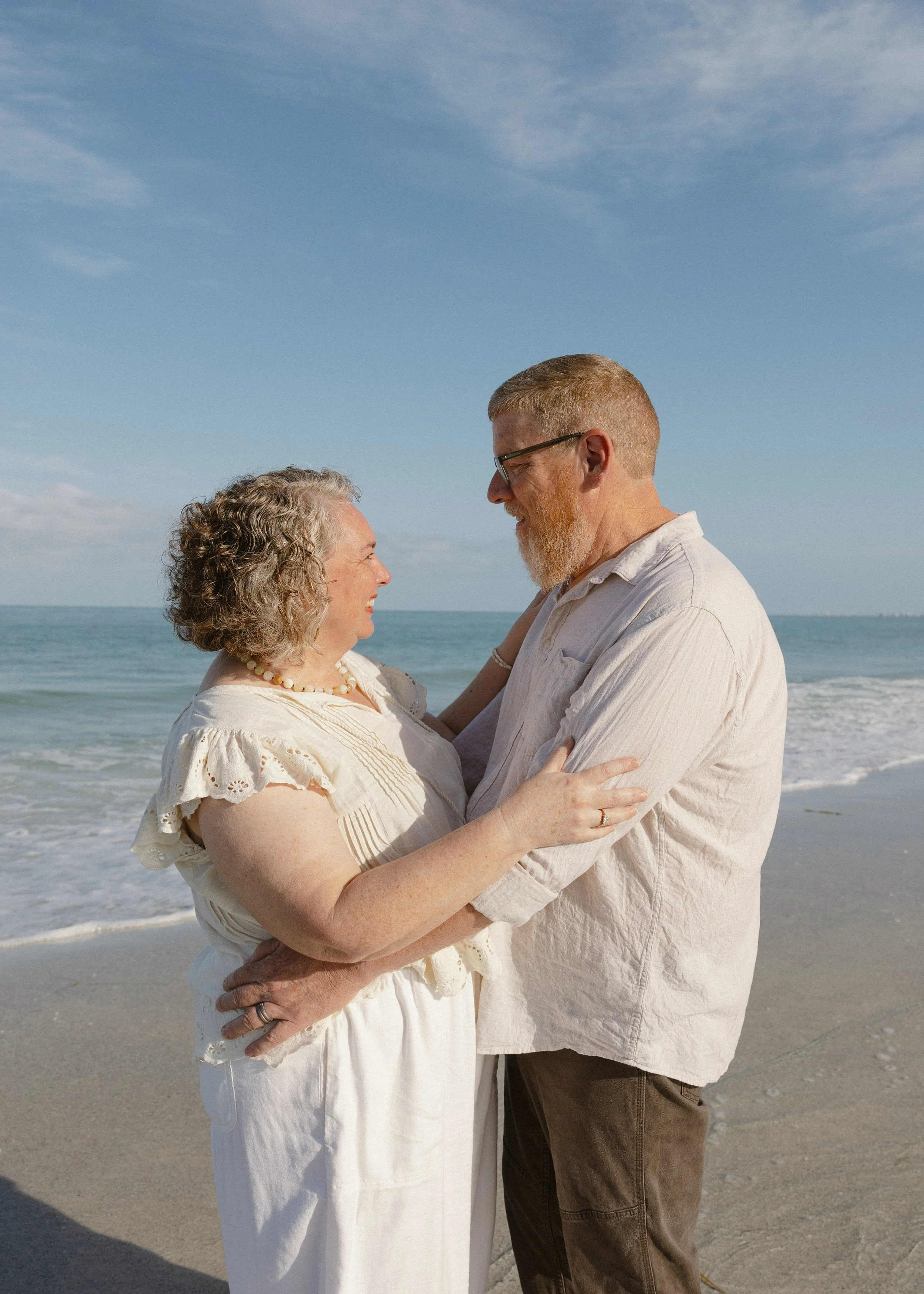 A middle-aged woman and a middle-aged man embrace and face each other on the beach, with the ocean and blue sky in the background.