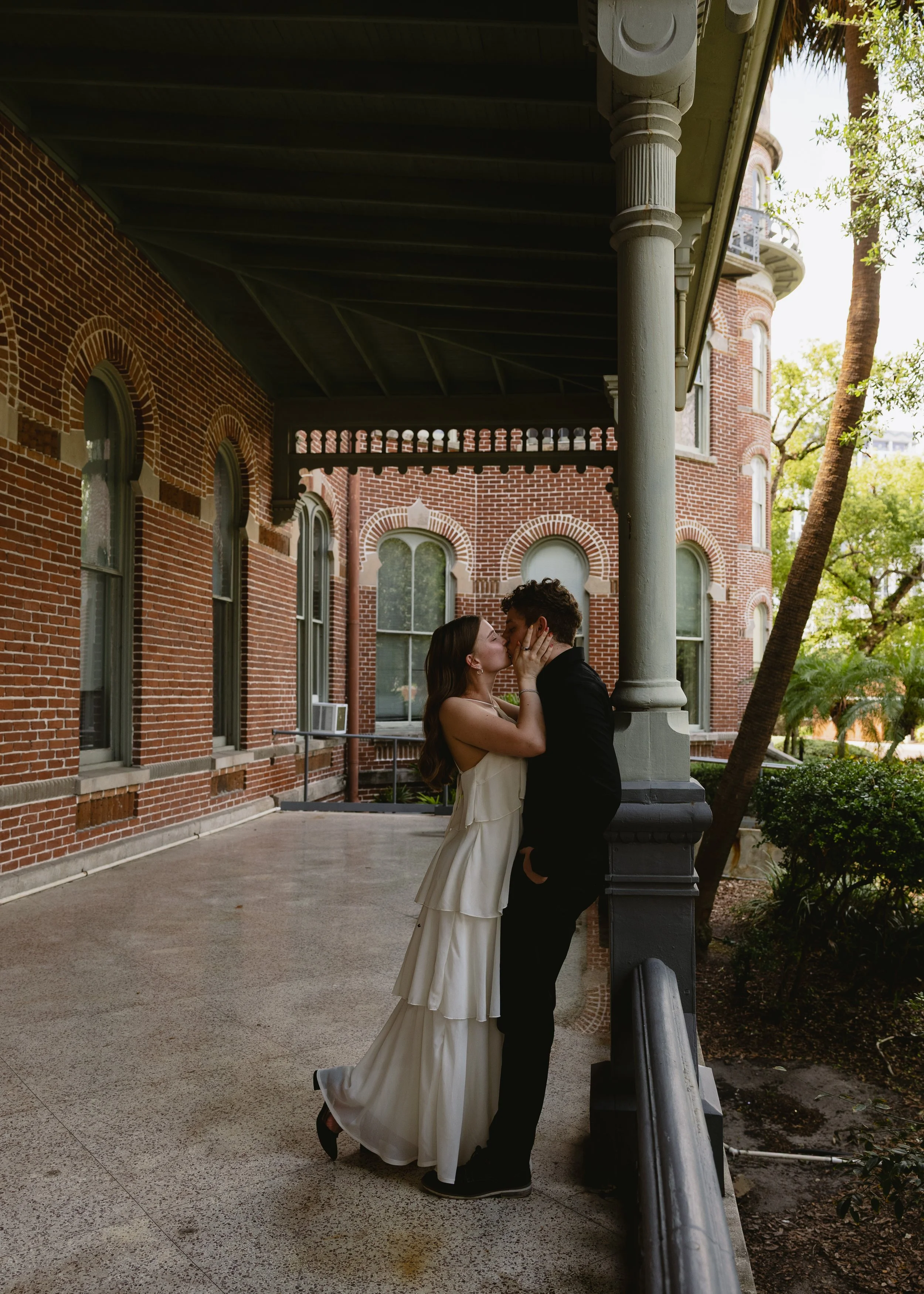 A man and woman kiss on a city sidewalk in front of a historic brick building with arched windows, surrounded by trees.
