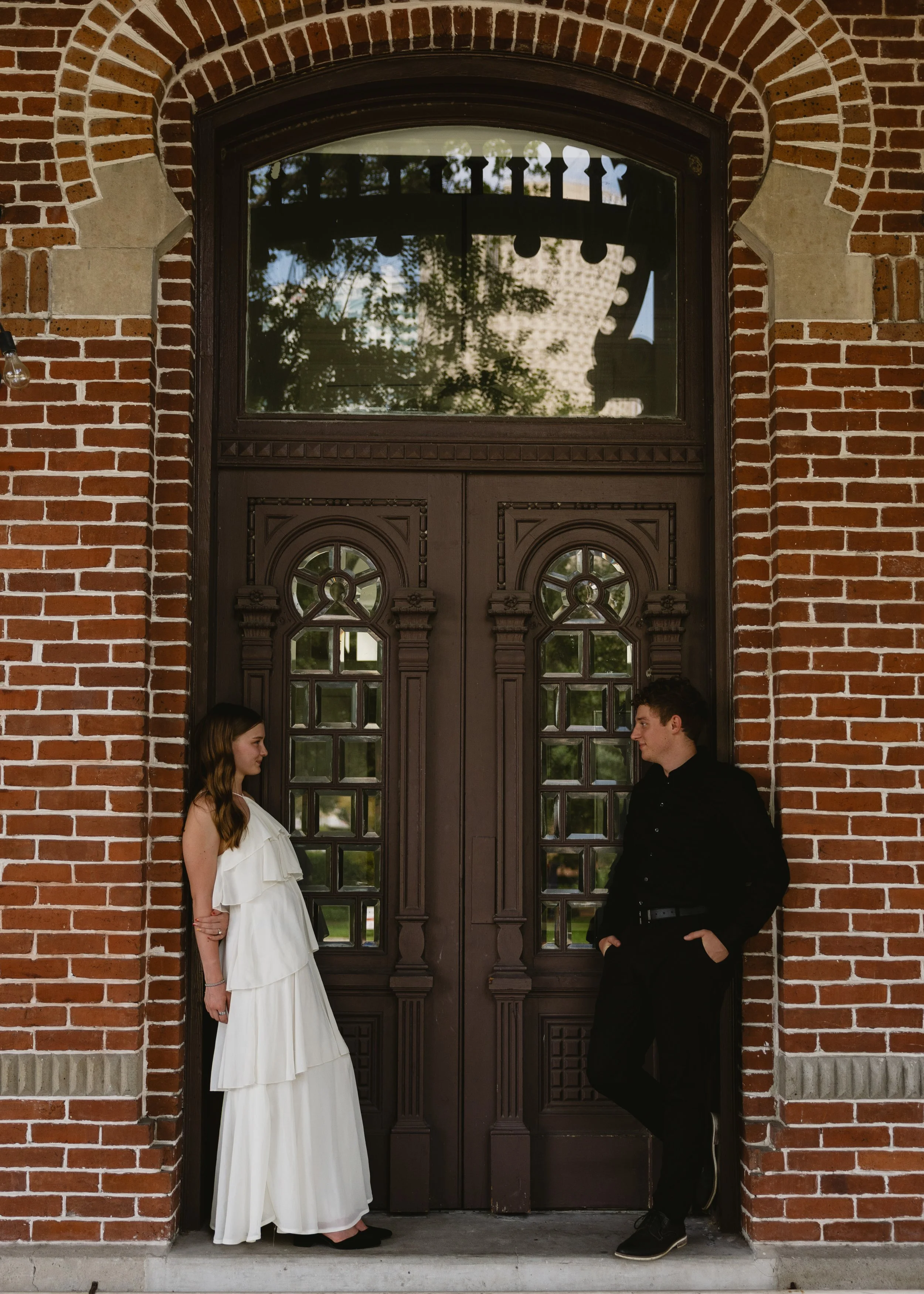 A woman in a white dress and a man in black clothing stand facing each other on either side of an ornate wooden door with a brick archway. They are positioned outdoors during daytime, with trees reflected in the door's glass.