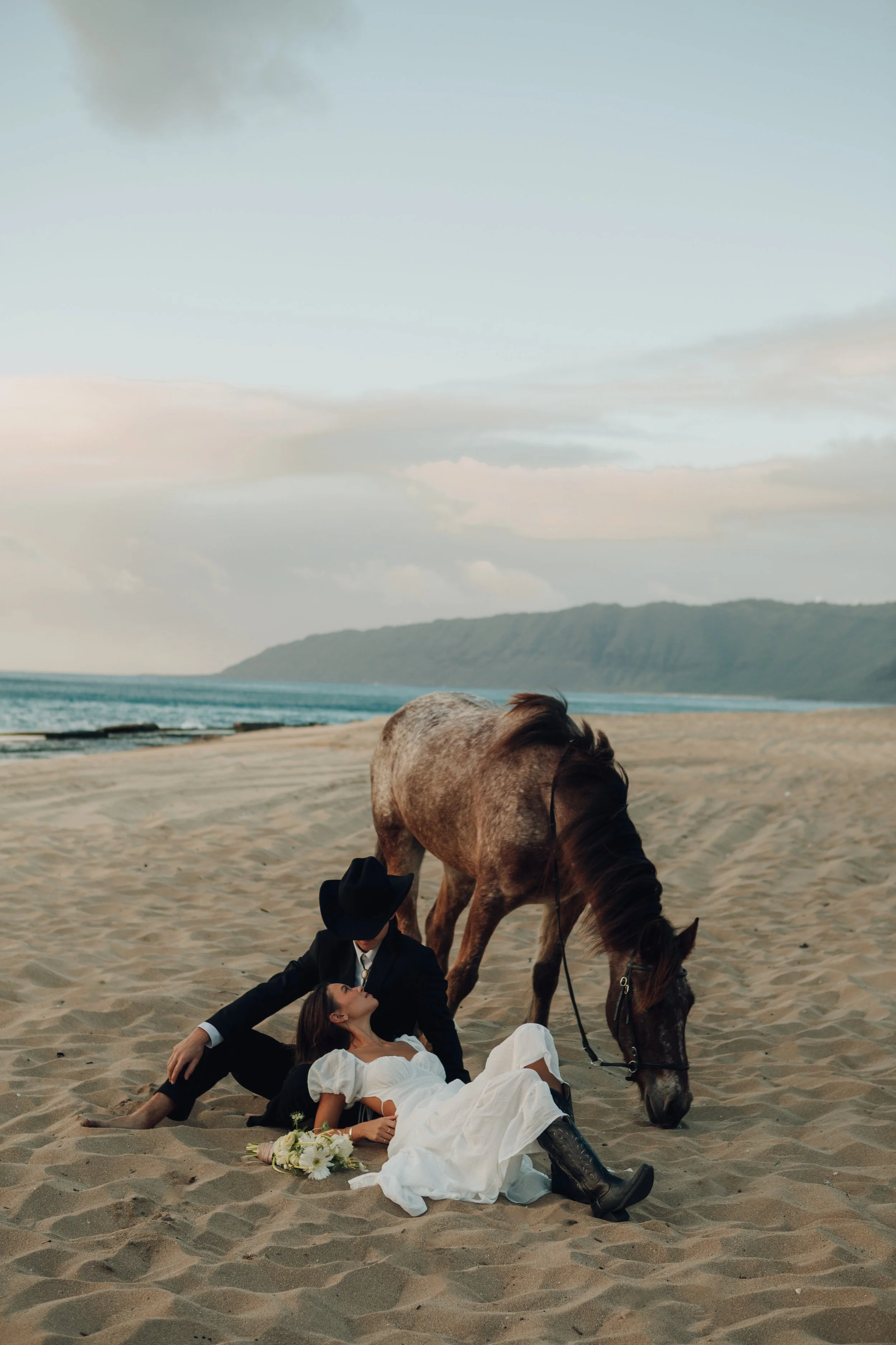 A couple lying on the sandy beach with a horse grazing above them, holding the woman's hand. The woman is dressed in a white wedding gown, and the man in a black suit and cowboy hat. Flowers are placed near the woman. In the background, the ocean wit