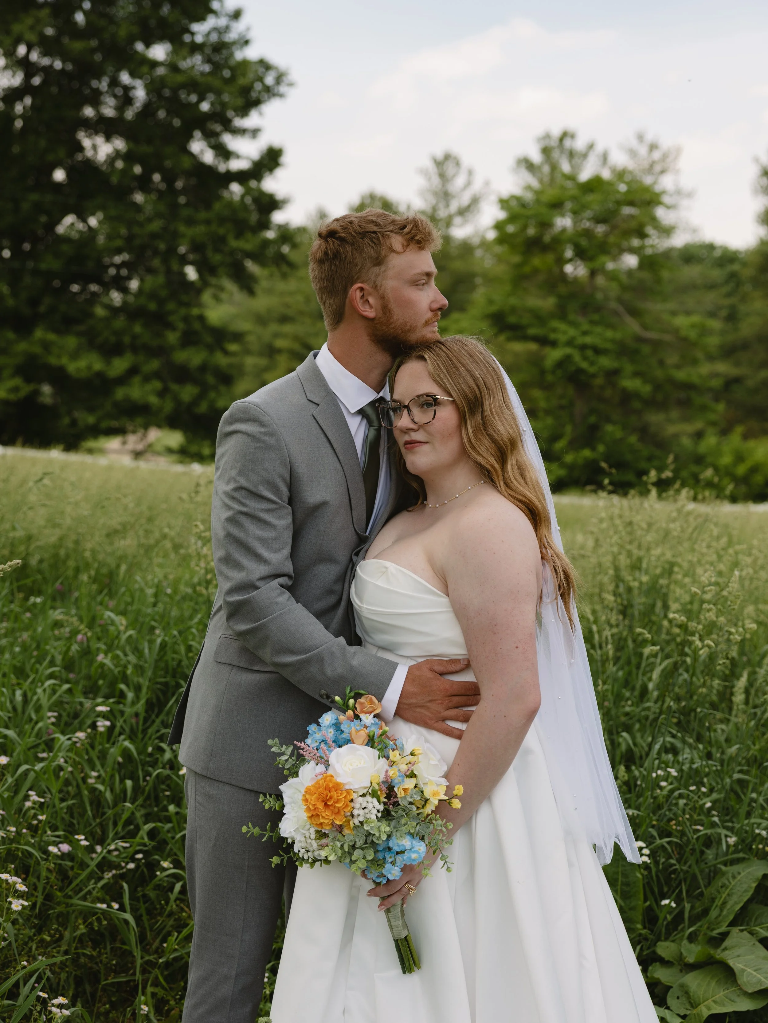 A newlywed couple standing in a grassy outdoor area surrounded by trees. The groom, dressed in a gray suit and white shirt, has red hair and a beard. The bride, with long red hair and glasses, wears a strapless white wedding dress and holds a bouquet