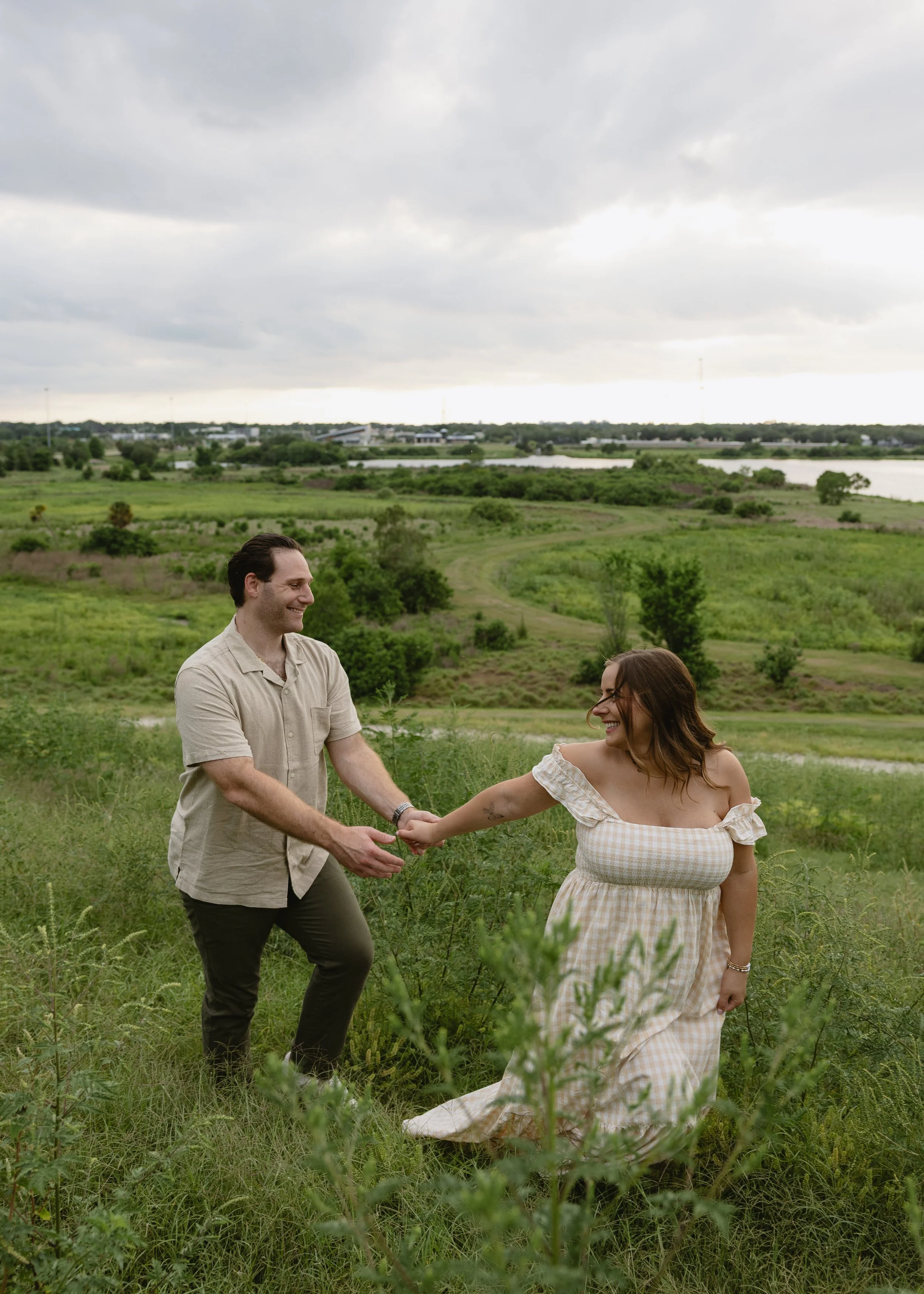 A man and woman holding hands in a green field with overcast sky, smiling and looking at each other.